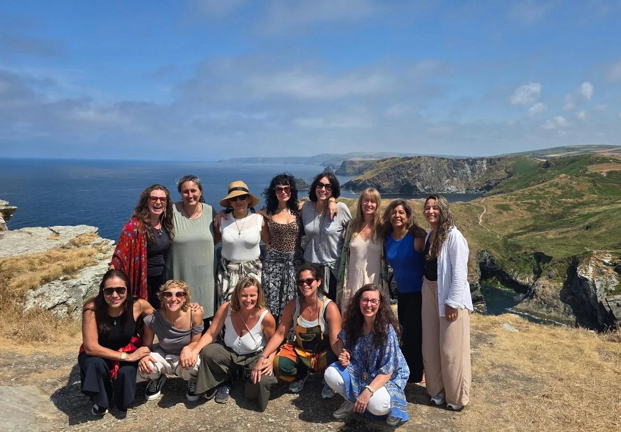 Grupo de quince mujeres posando en un paisaje costero con acantilados y mar azulado en el fondo, con cielo parcialmente nublado.