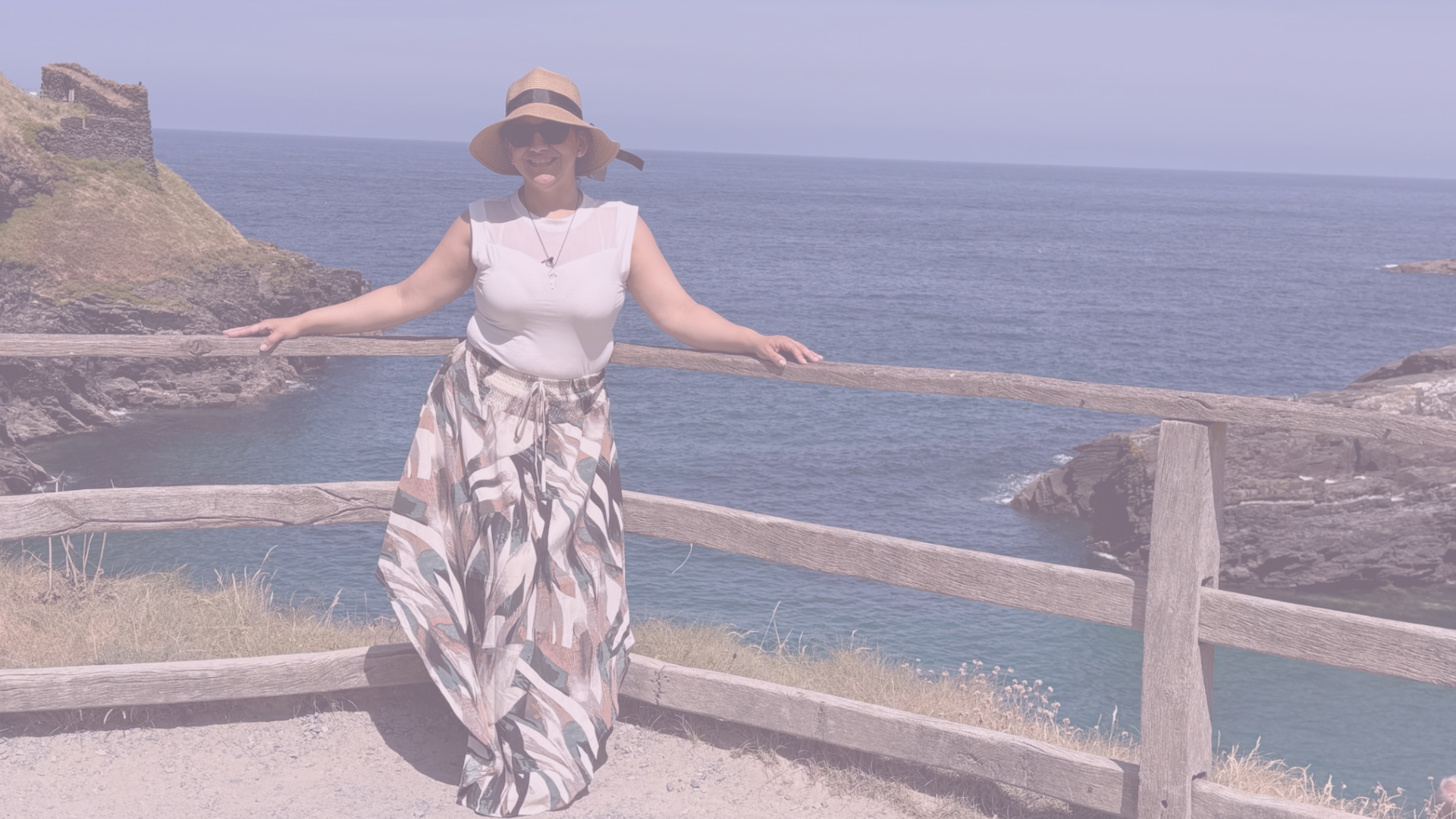 Mujer con sombrero y gafas de sol posando en un mirador con vista al océano y un castillo en la colina.