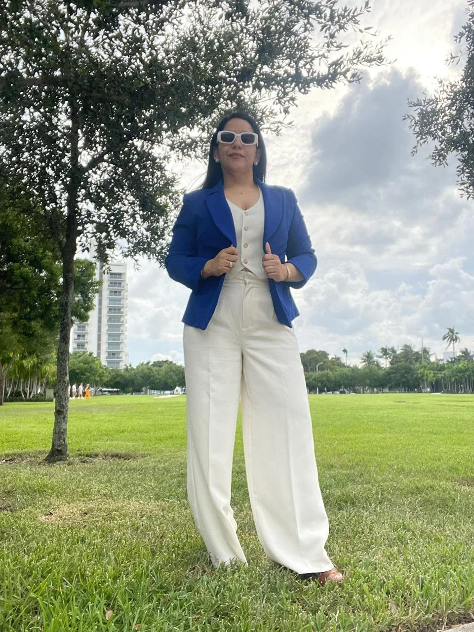 Una mujer de pie en un parque con césped verde, viste un traje azul y pantalones blancos, lleva gafas blancas, y detrás hay árboles, un edificio alto y un cielo con nubes.