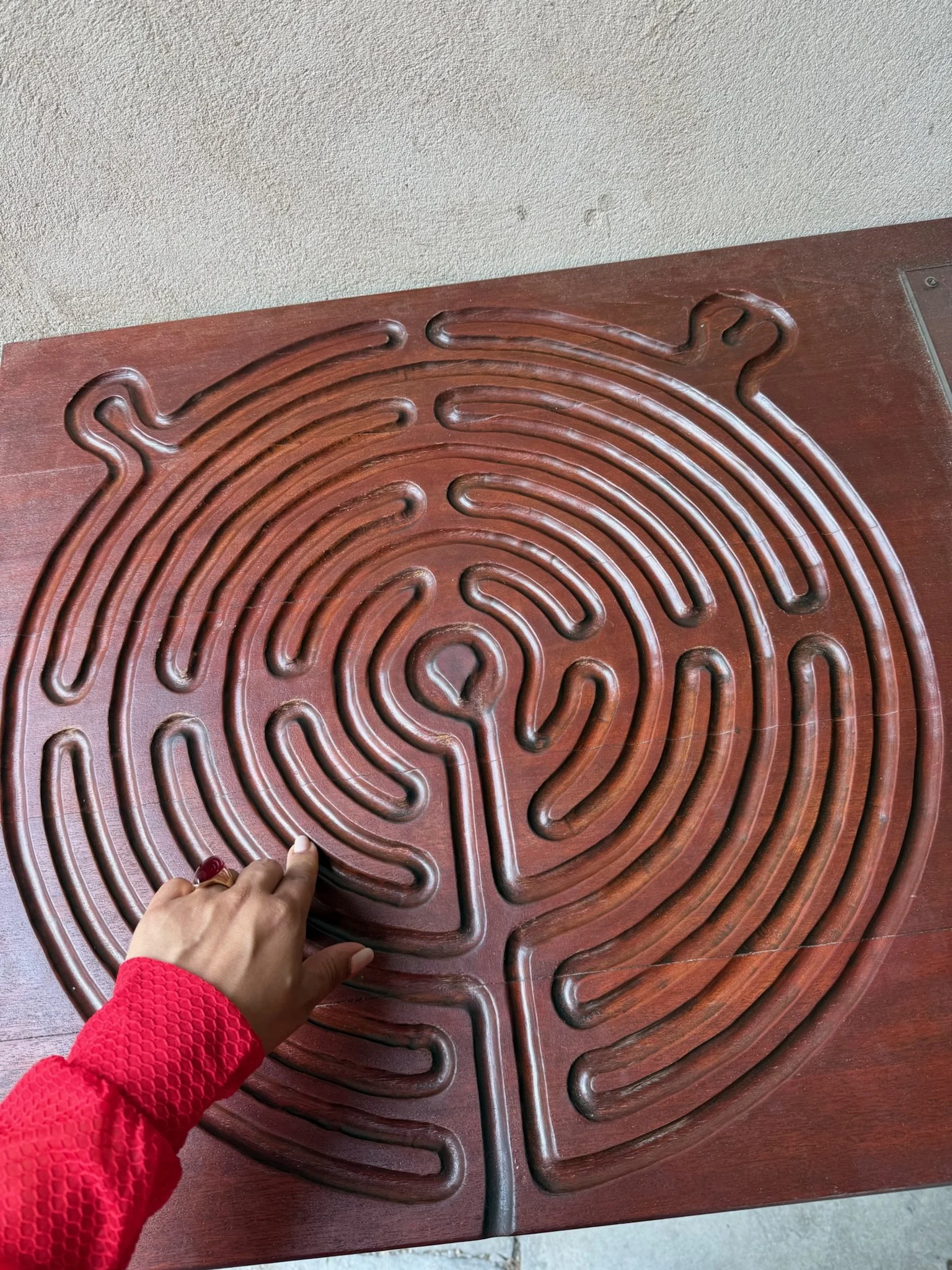 Persona apuntando con un dedo una cerradura en forma de laberinto en una mesa de madera, usando un suéter rojo