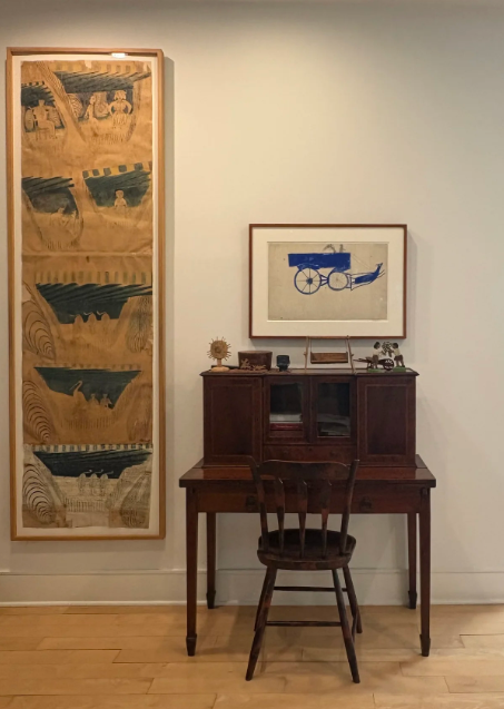 A vintage wooden desk with a matching chair set against a white wall, with framed artwork hanging above. To the left, a tall vertical scroll with a tan background and black and blue artwork.