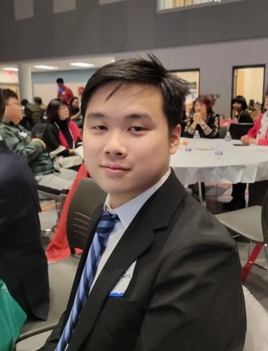 A young man in a suit and tie sitting at a table in a crowded indoor event or conference.