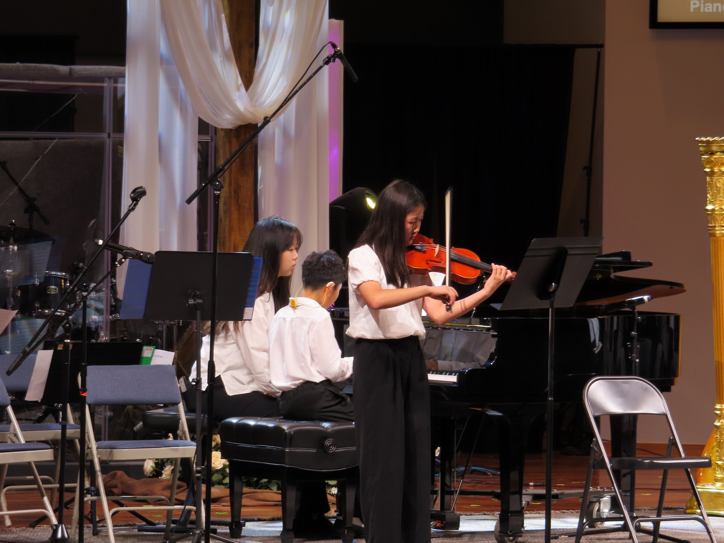 Young woman with long hair braided on the side, sitting at a black grand piano on stage, wearing a white shirt and black pants, with a purple-lit stage background.
