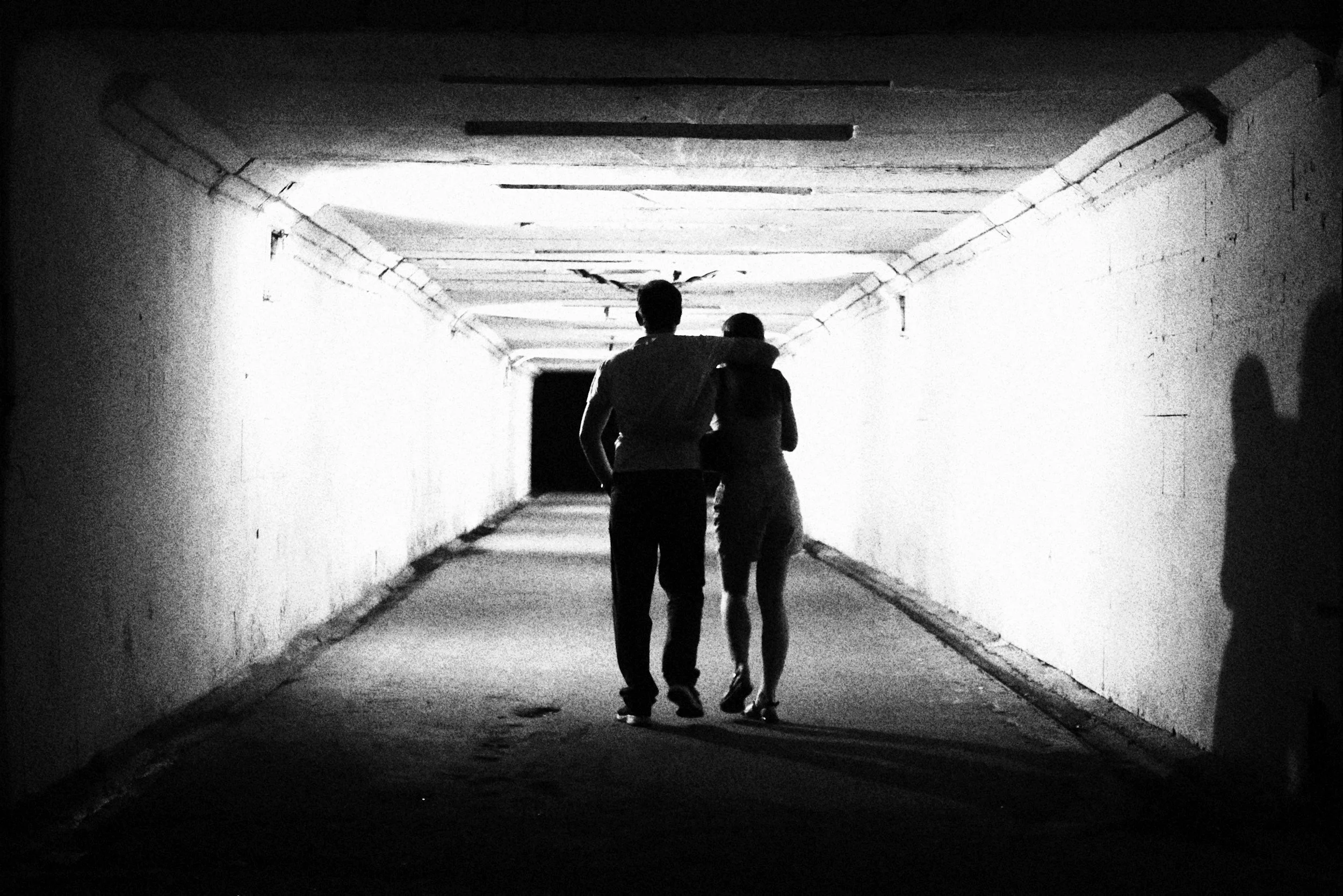 A black and white photo of two people walking through a dimly lit underpass or tunnel, with one person having their arm around the other's shoulders.