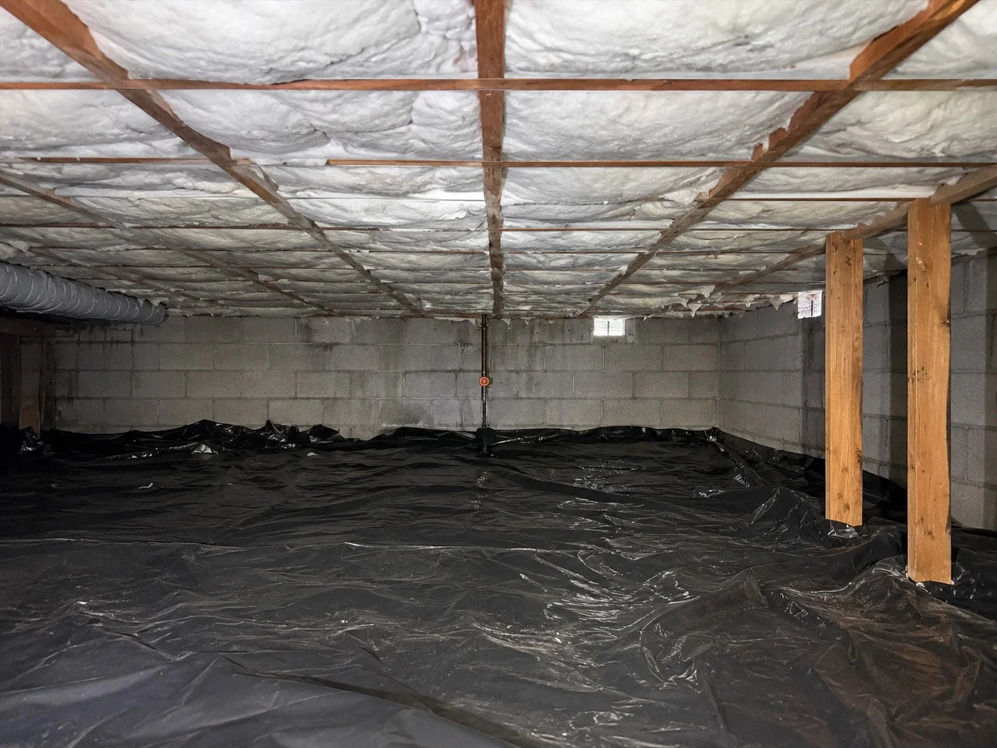 Crawlspace with batting insulation in floor, a concrete block wall, a black vapor barrier on the floor, and wooden support beams.