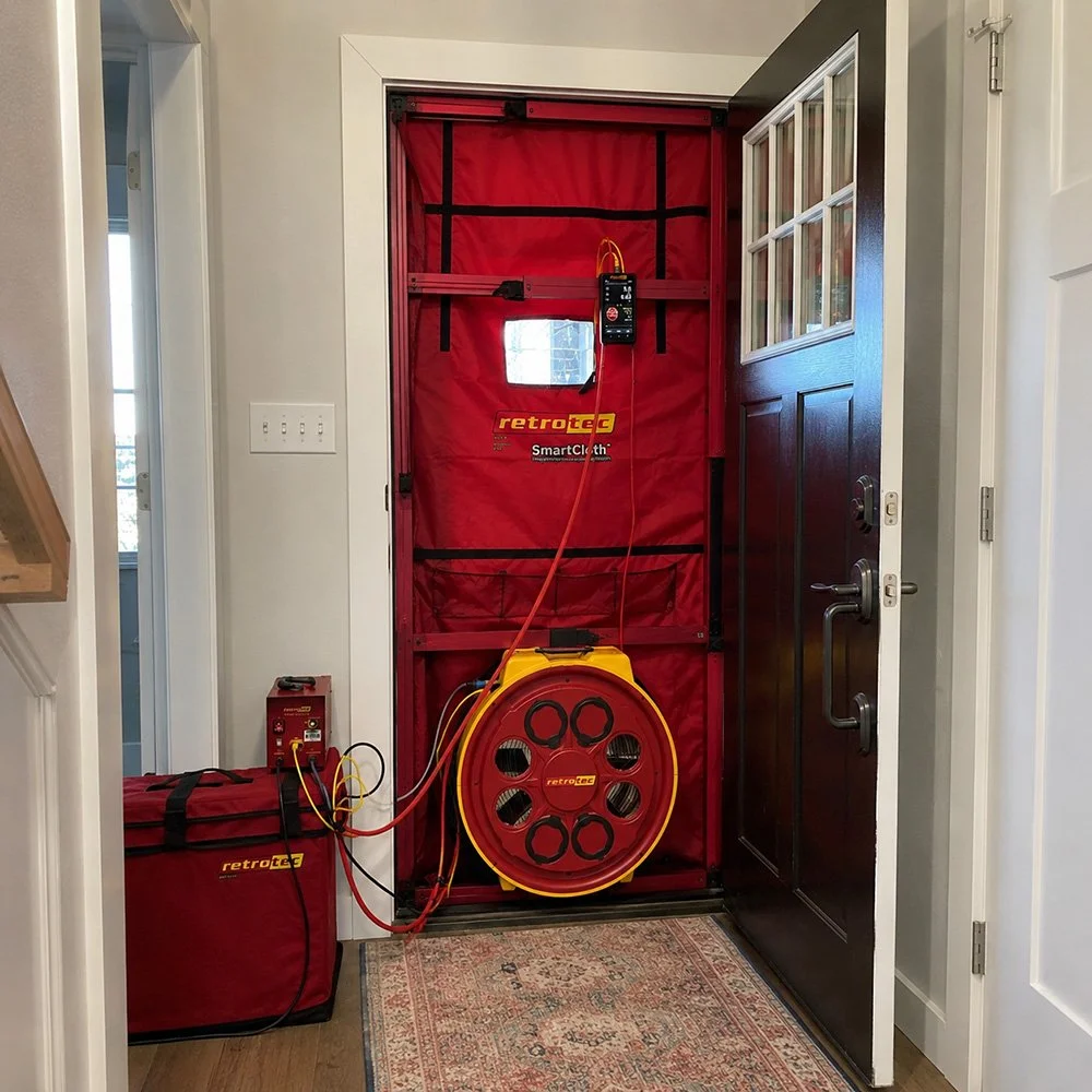 A blower door testing system with a red fabric cover, connected cables, and testing equipment, set up at a doorway.