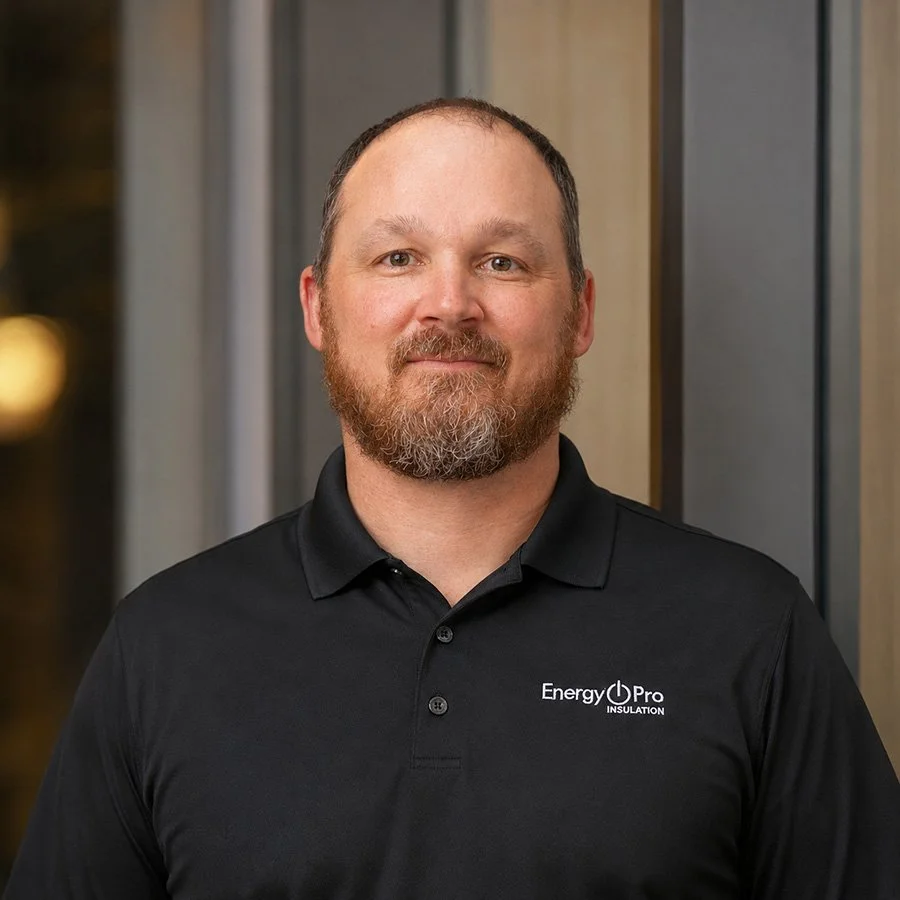 A man with a beard and short hair wearing a black polo shirt with a logo that reads 'Energy Pro Insulation,' standing in front of a modern wooden and glass wall.