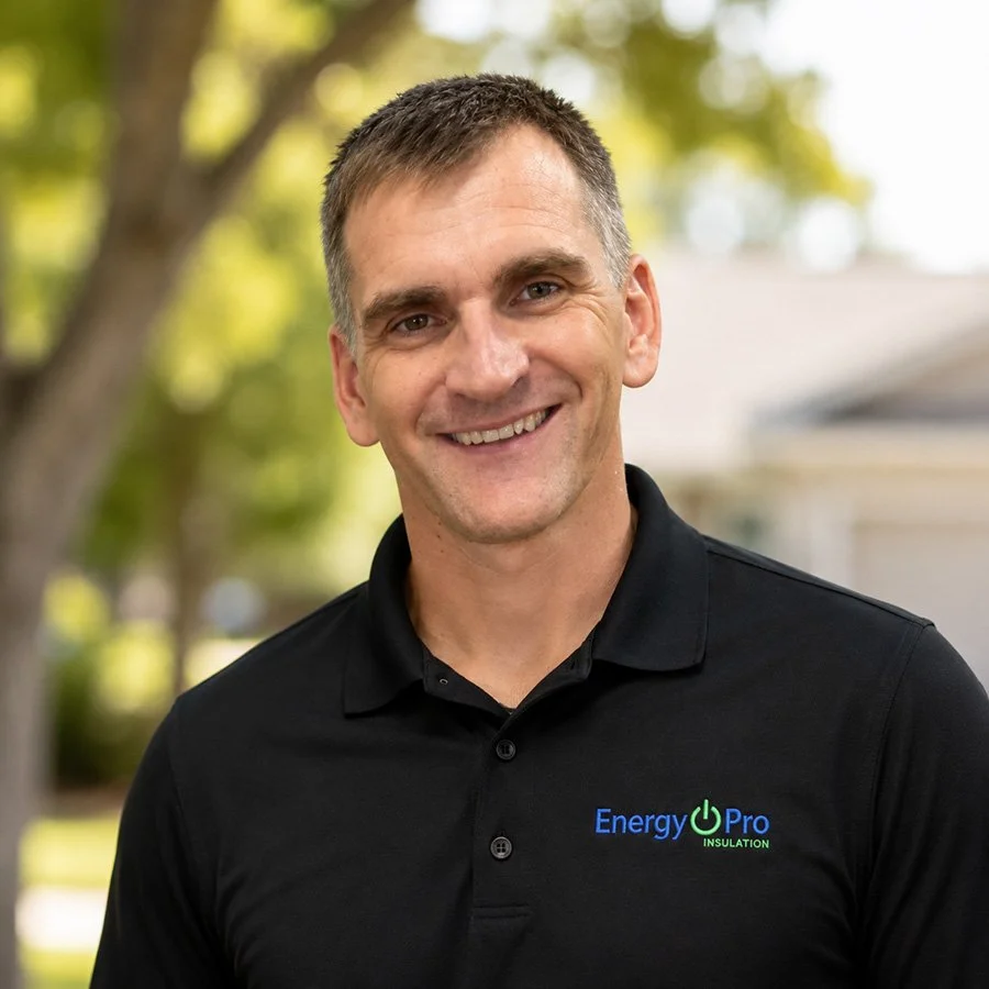 Smiling man outdoors wearing a black polo shirt with 'Energy Pro Insulation' logo, blurred trees and house in background.