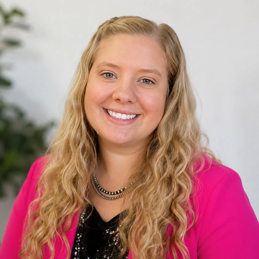 A young woman with long, wavy blonde hair, smiling, wearing a pink blazer and black top with sequins, and a black-and-silver beaded necklace.