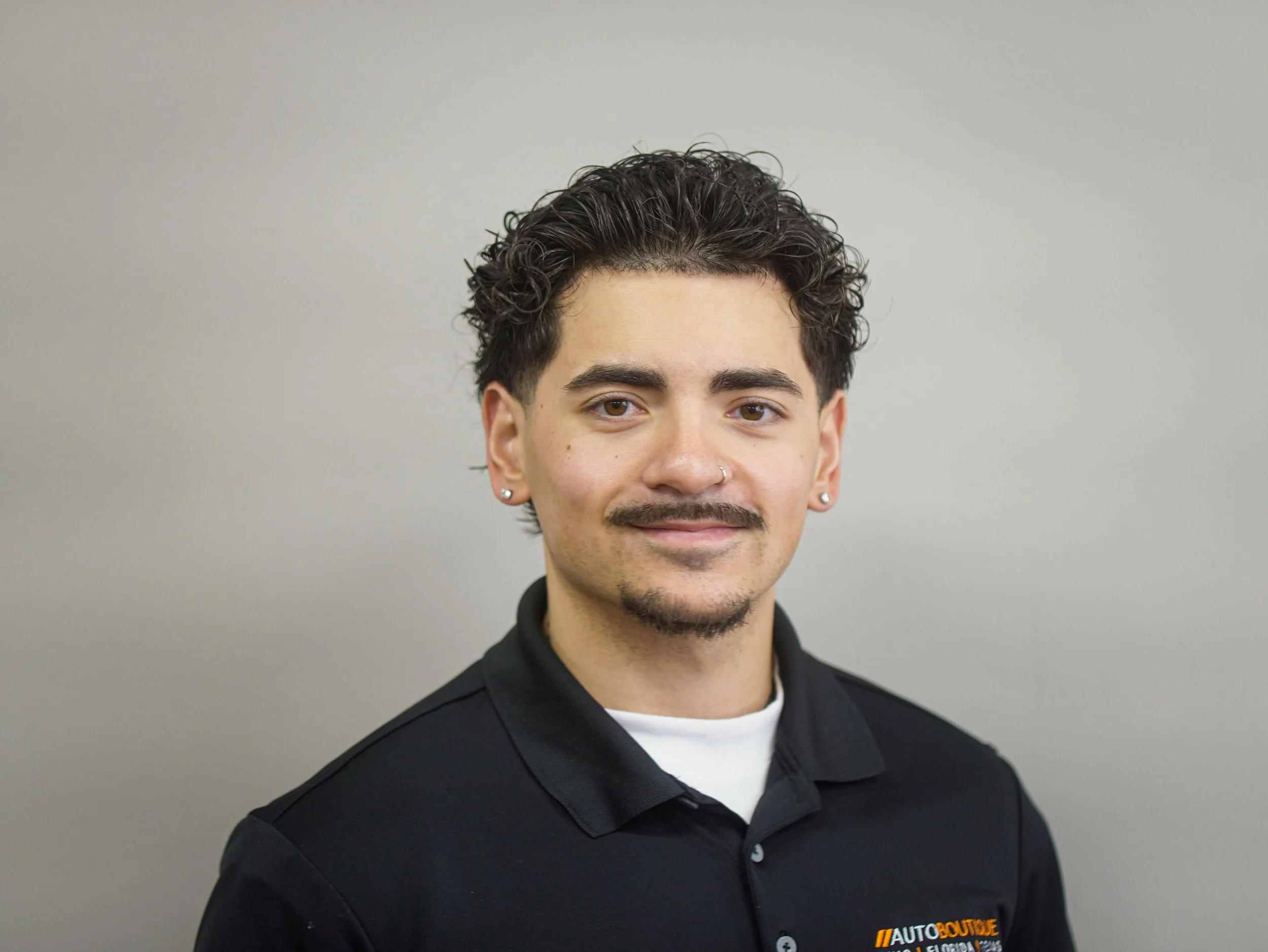 Portrait of Tavian with dark curly hair, light skin, wearing earrings, a nose ring, and a black collared shirt with a small logo that says 'Auto Boutique' in orange and white on the left chest, standing against a plain gray background.