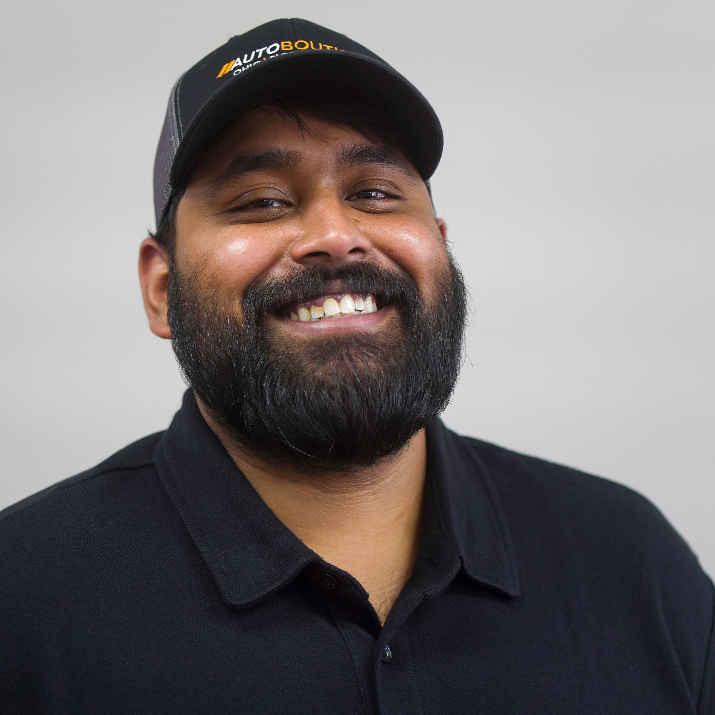 A portrait of Andrew with a dark beard and mustache, smiling and wearing a black polo shirt and a black cap with 'AUTO BOUTIQUE' logo, standing against a plain light gray background.