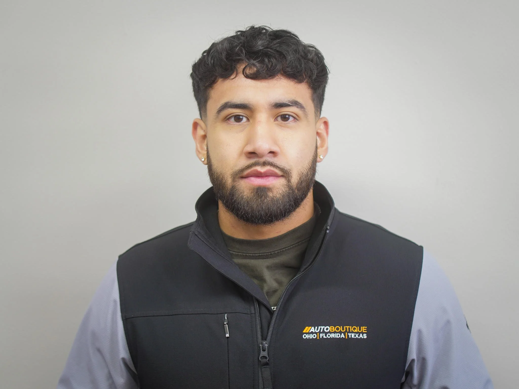 A young man with a beard and earrings looking directly at the camera, wearing a black and gray zip-up jacket with a logo for Auto Boutique.