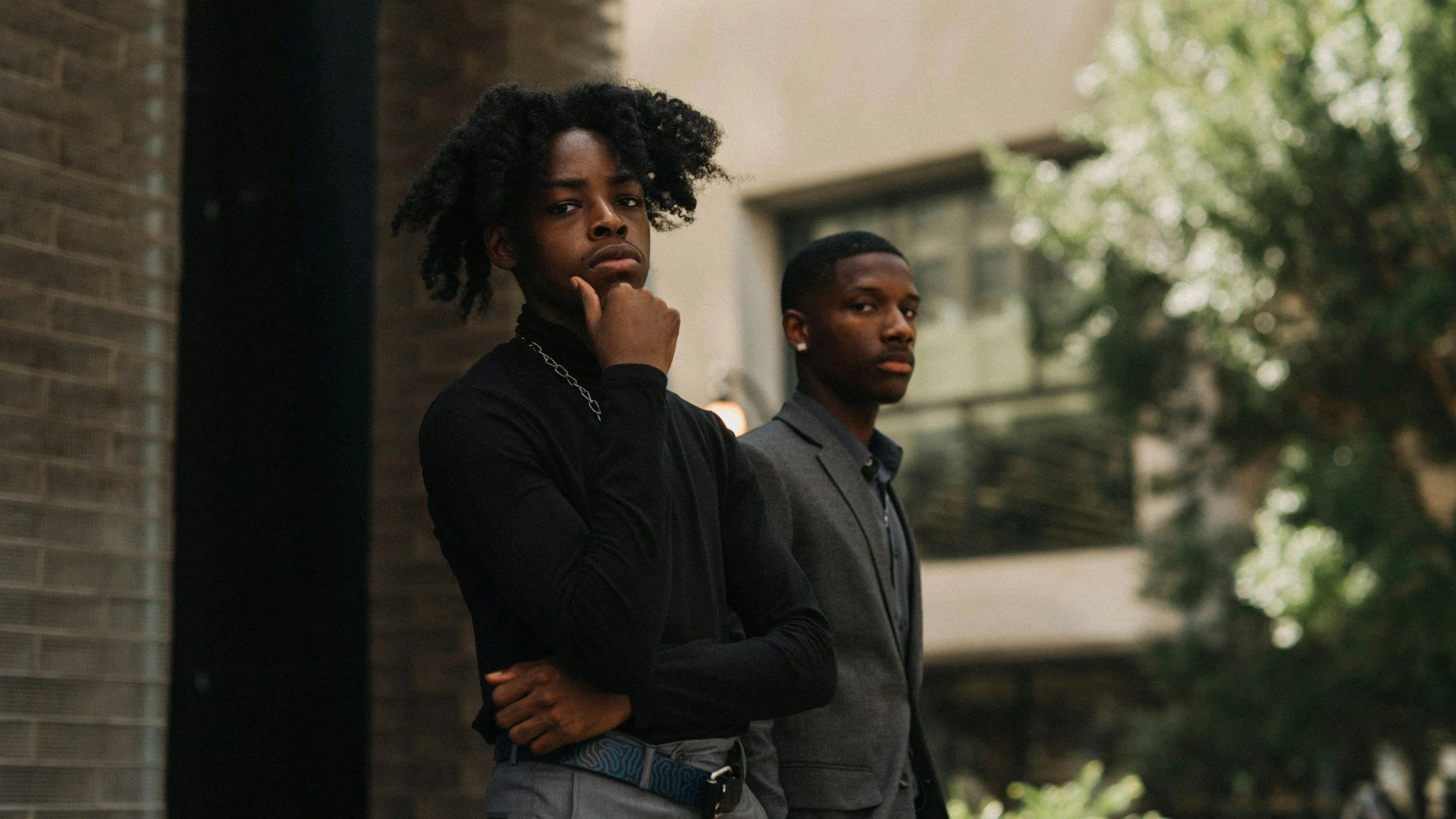 Two young men standing outdoors, one with hand on chin and the other with arms crossed, near a building with trees in the background.