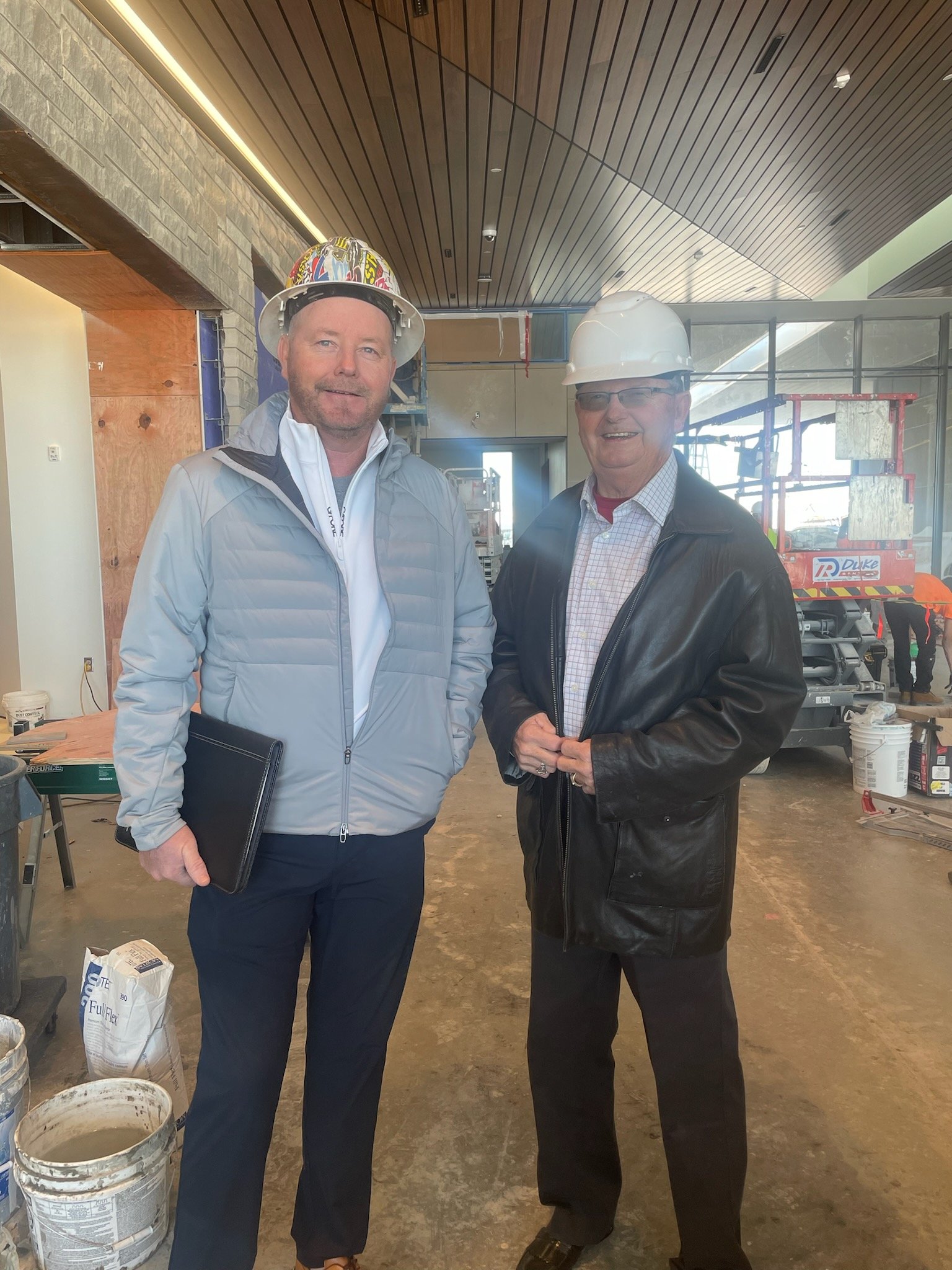 Two men wearing hard hats standing inside a building under construction. One is holding a folder, and construction equipment and materials are visible in the background.