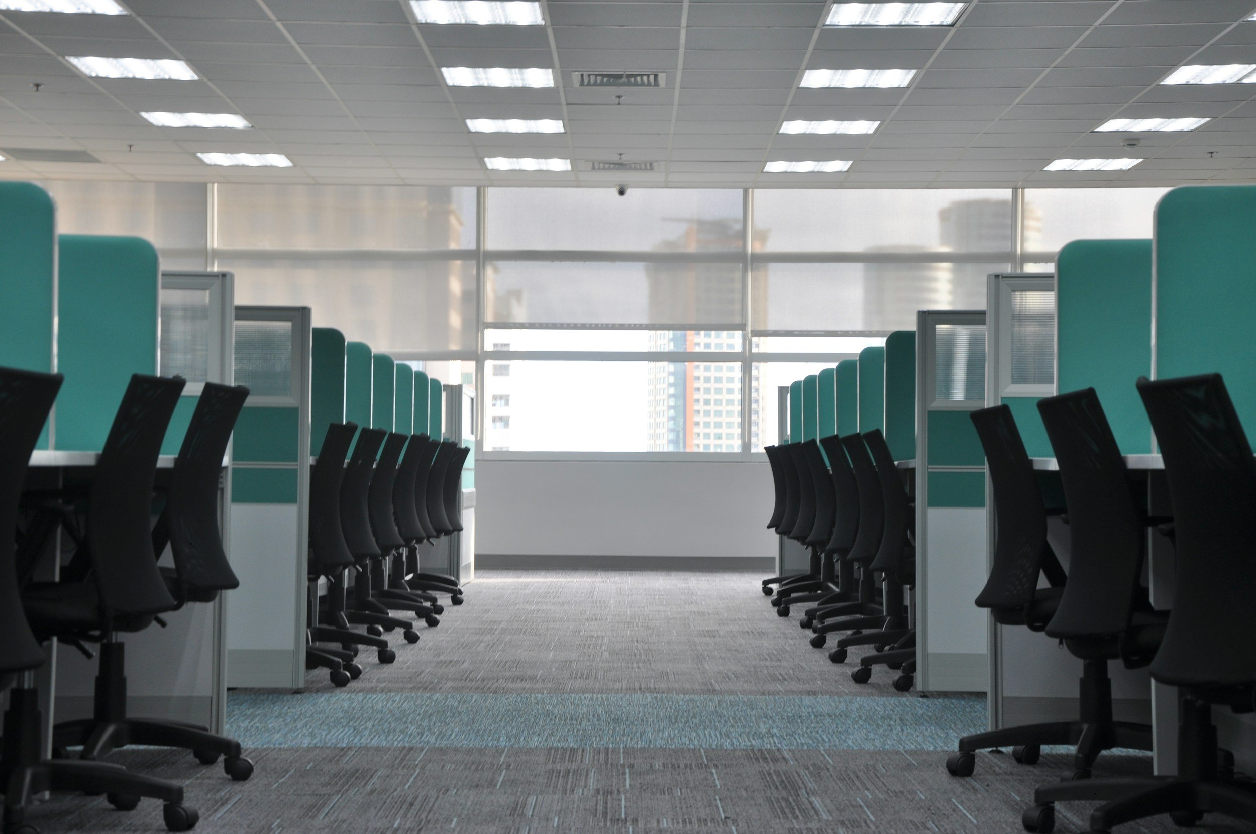 Empty office cubicles with black chairs, teal dividers, and large windows showing city buildings.