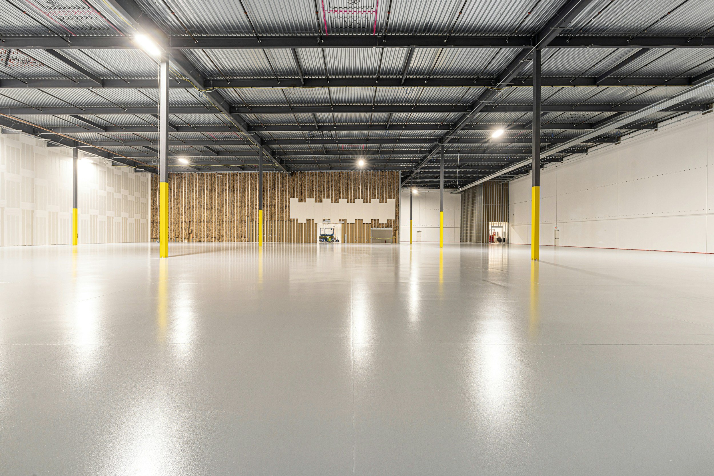 Empty warehouse with polished concrete floor, black ceiling with fluorescent lights, and yellow safety posts.