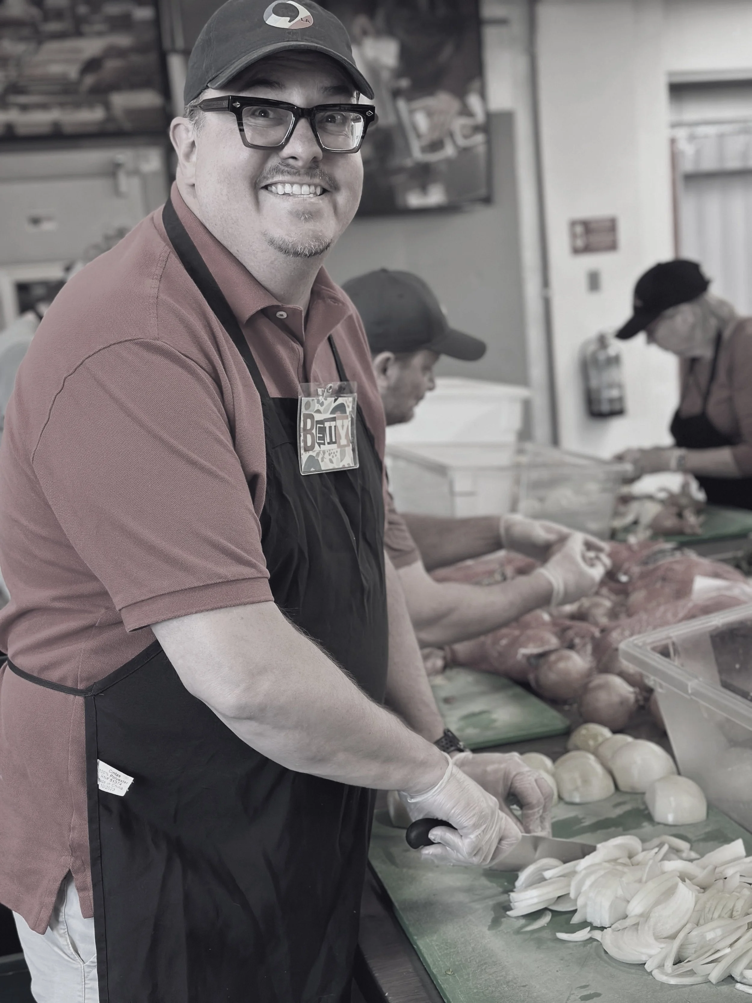 A man wearing glasses, a cap, and an apron smiles while chopping onions in a kitchen or food prep area. In the background, other people are working with similar ingredients.