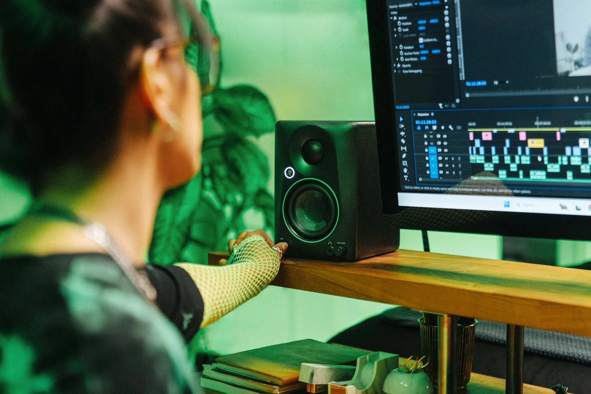 A person operating music production software on a computer with a monitor displaying multiple audio tracks and waveforms. The person is touching a studio monitor speaker on a wooden desk.