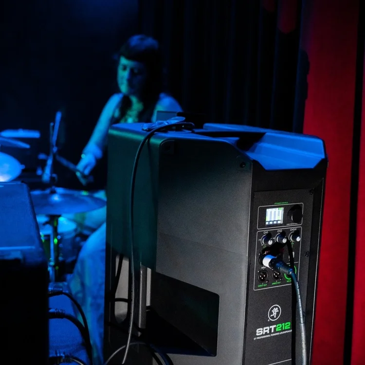 A woman playing drums on stage with blue and red lighting, with a large black speaker in the foreground.