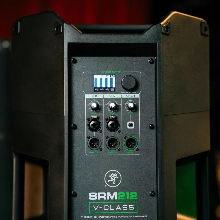 Front view of a black powered loudspeaker with control panel showing input ports, knobs, and a digital display