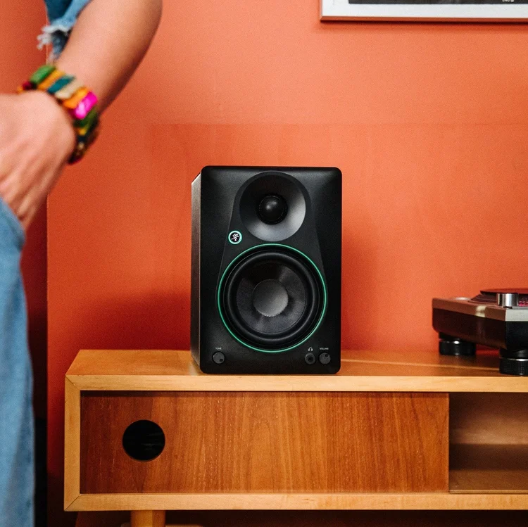 A black studio monitor speaker on a wooden table in front of a reddish-orange wall, with part of a person's arm and colorful bracelet visible on the left side.