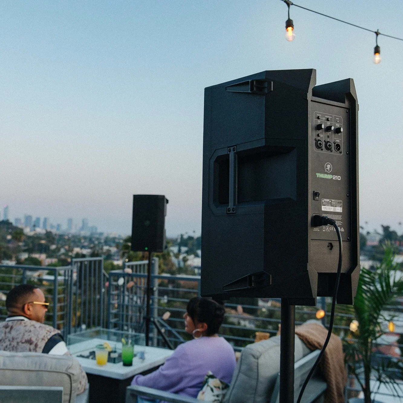 Outdoor rooftop gathering at sunset with two people seated, a speaker on a stand, string lights, and a city skyline in the background.