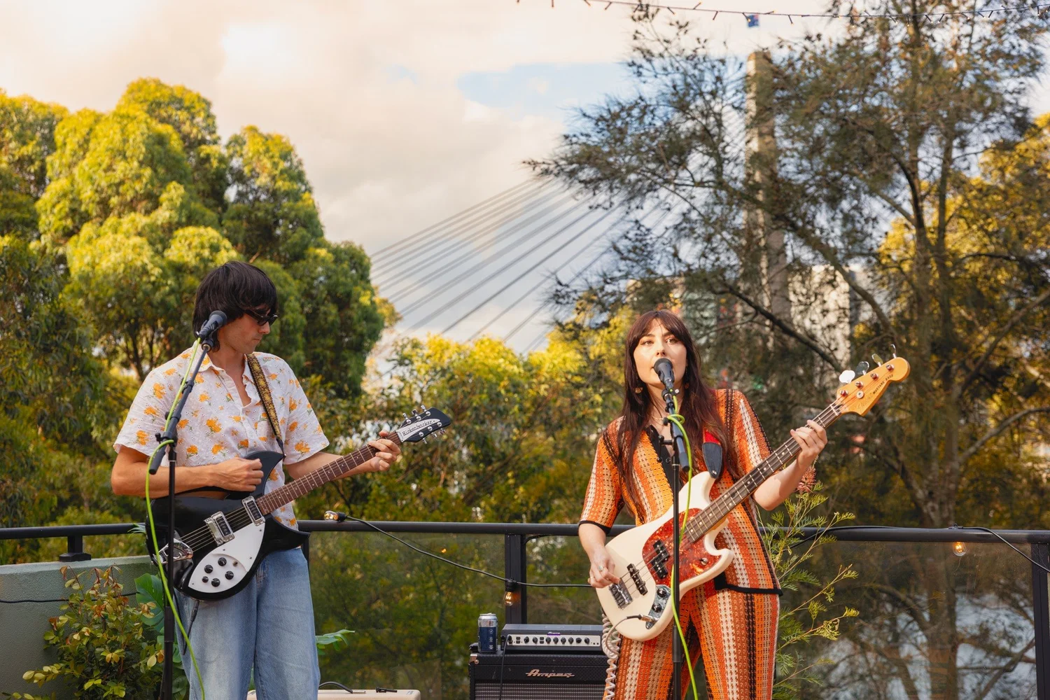 Two women performing music outdoors on stage, one playing guitar and the other playing bass guitar, with a backdrop of green trees and a partly cloudy sky.