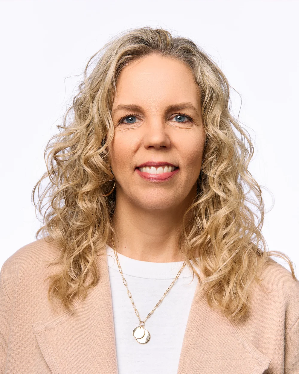 A woman with blonde curly hair, blue eyes, wearing a beige blazer, white top, and a gold necklace with layered circles, smiling at the camera against a plain white background.