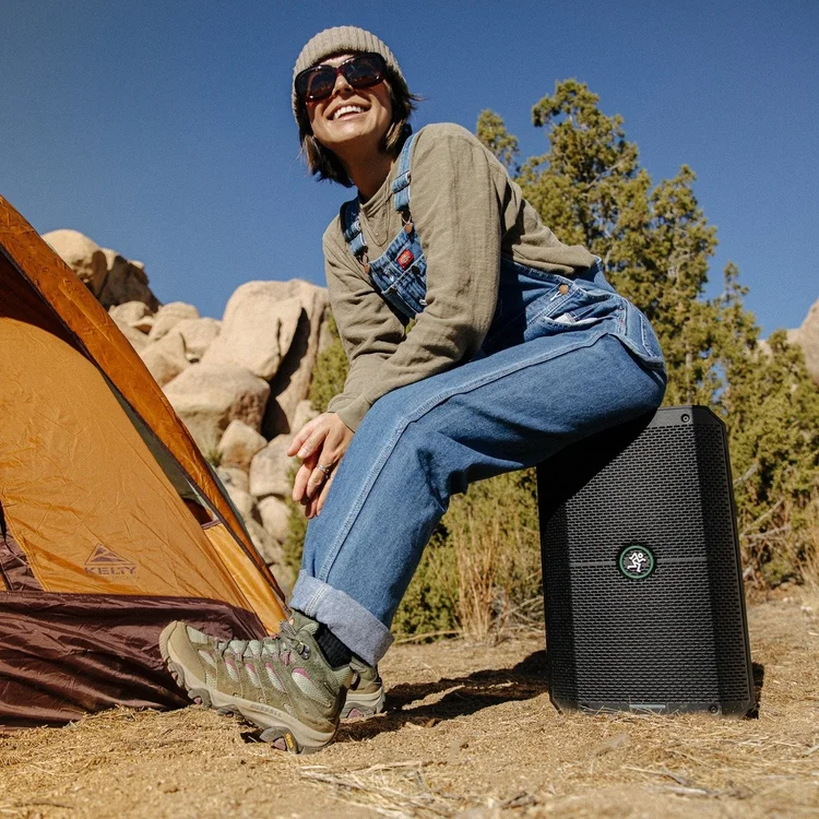 A woman wearing sunglasses, a beanie, and casual outdoor clothing sitting on a speaker near a camping tent outdoors with rocks and trees in the background under a clear blue sky.