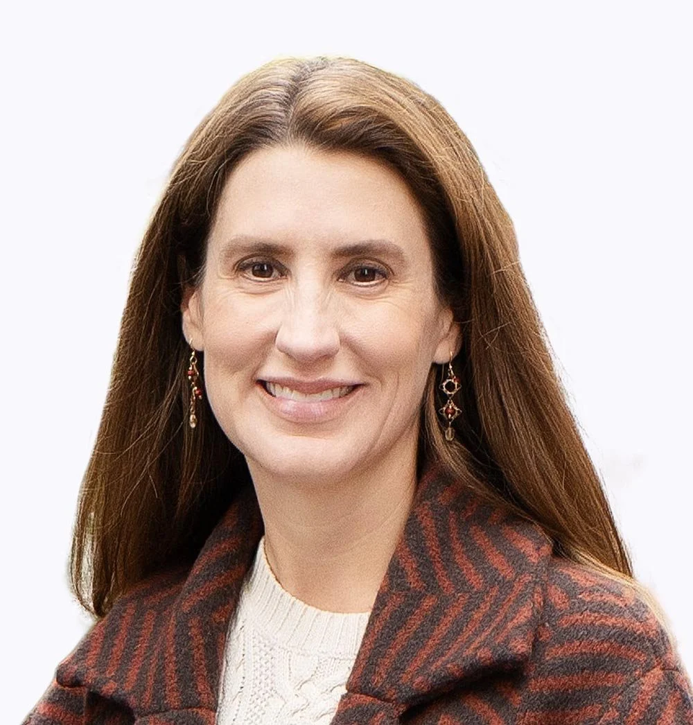 A woman with long brown hair smiling, wearing earrings, a plaid jacket, and a light-colored sweater against a plain background.