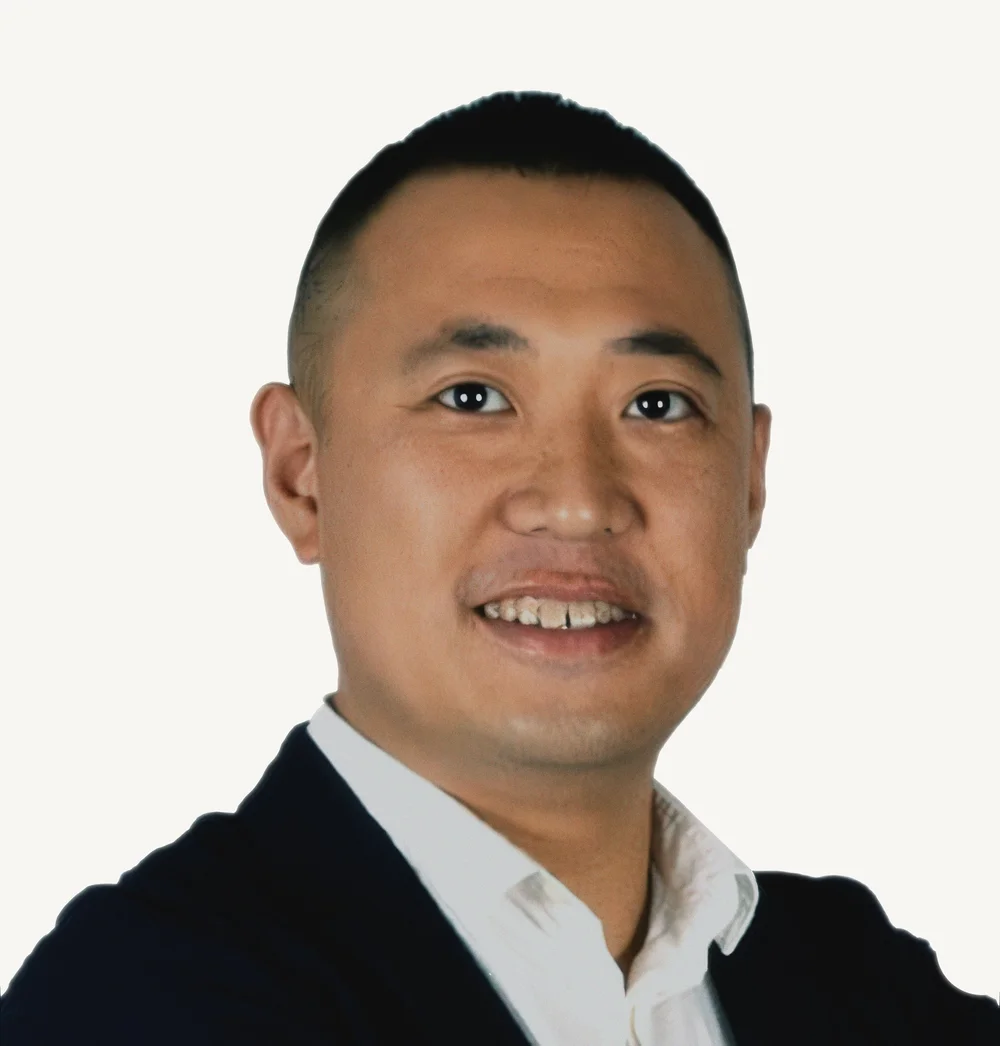 Headshot of a man with short black hair, wearing a black blazer over a white shirt, smiling at the camera against a plain white background.
