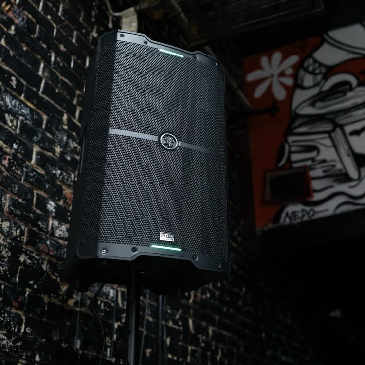 Black professional speaker mounted on a stand in front of a dark brick wall with graffiti artwork