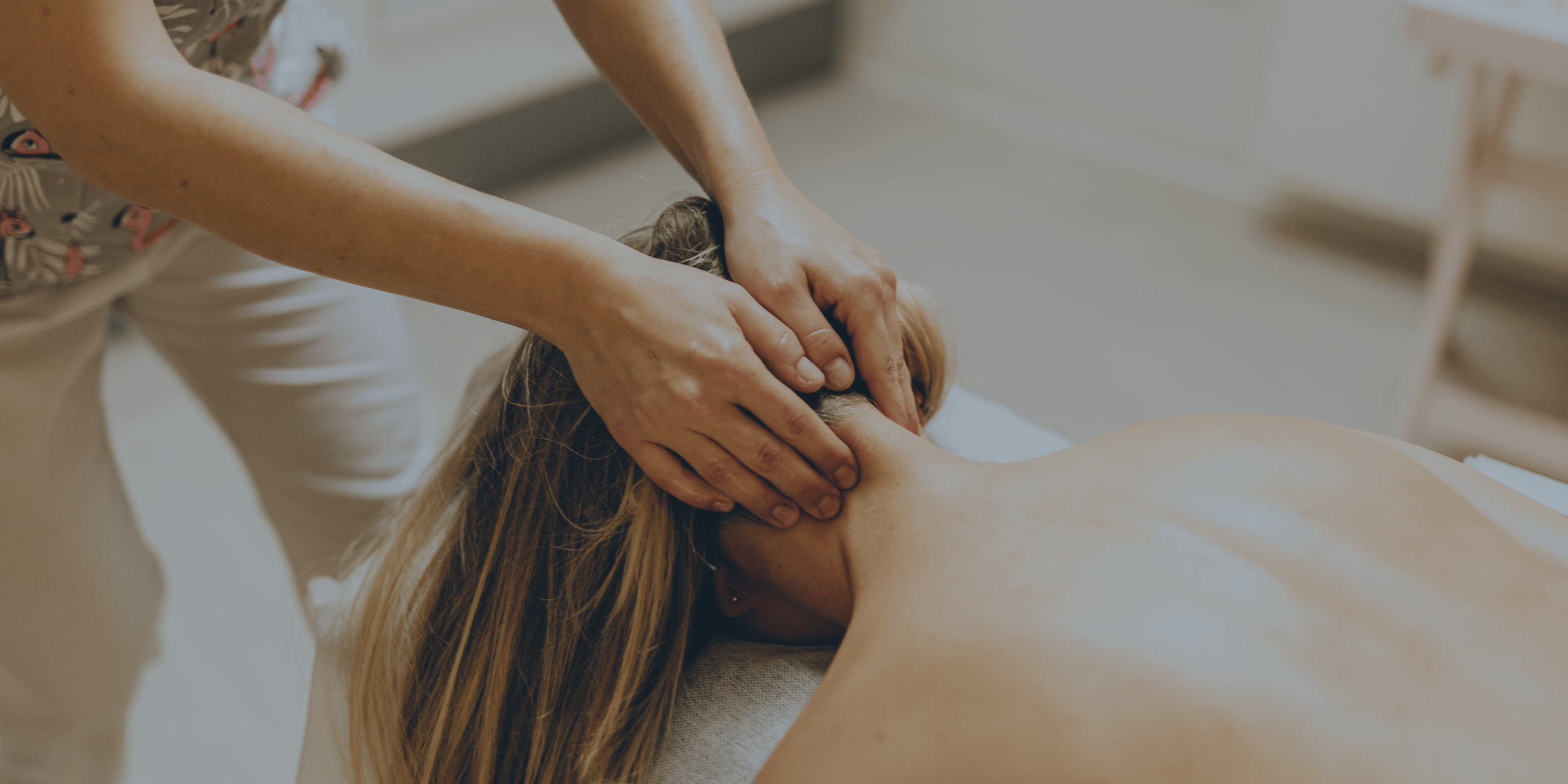 Close-up of a massage therapist's hands applying massage on a person's back, lying face down on a massage table, in a well-lit room with wooden floor and furniture.