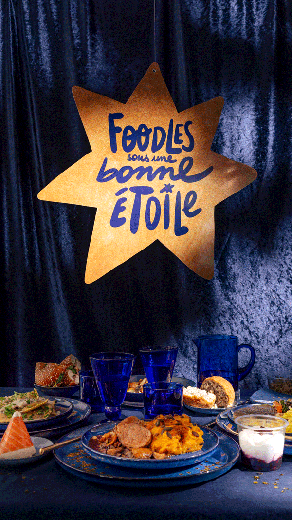 Table avec plats variés, desserts, verres bleus et une pancarte en forme d'étoile portant le texte "Foodles sous une bonne étoile" contre un fond sombre.