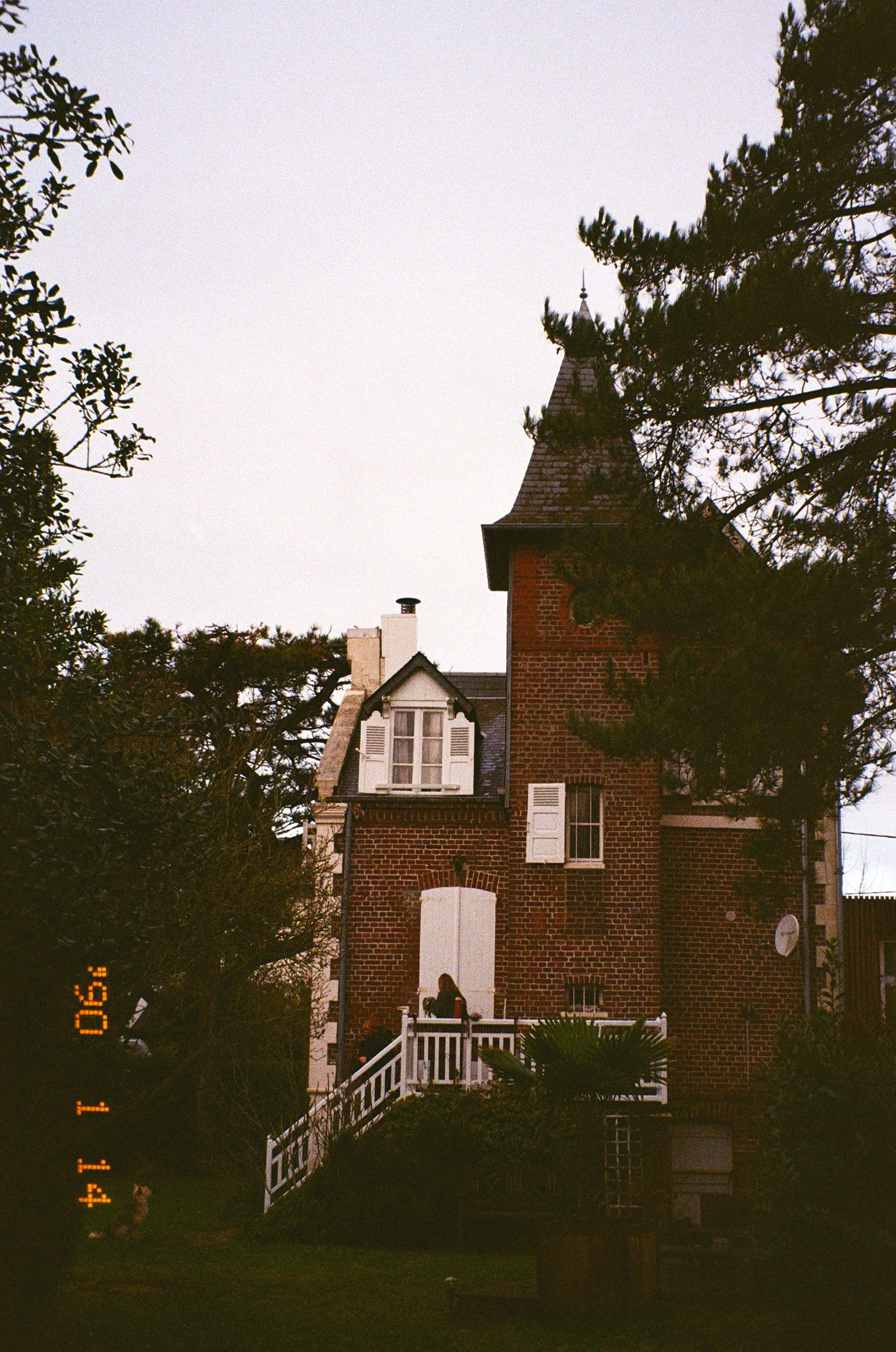 Une maison en briques rouges avec des fenêtres à volets blancs, un escalier menant à une petite terrasse, entourée d'arbres et de plantes, sous un ciel clair.