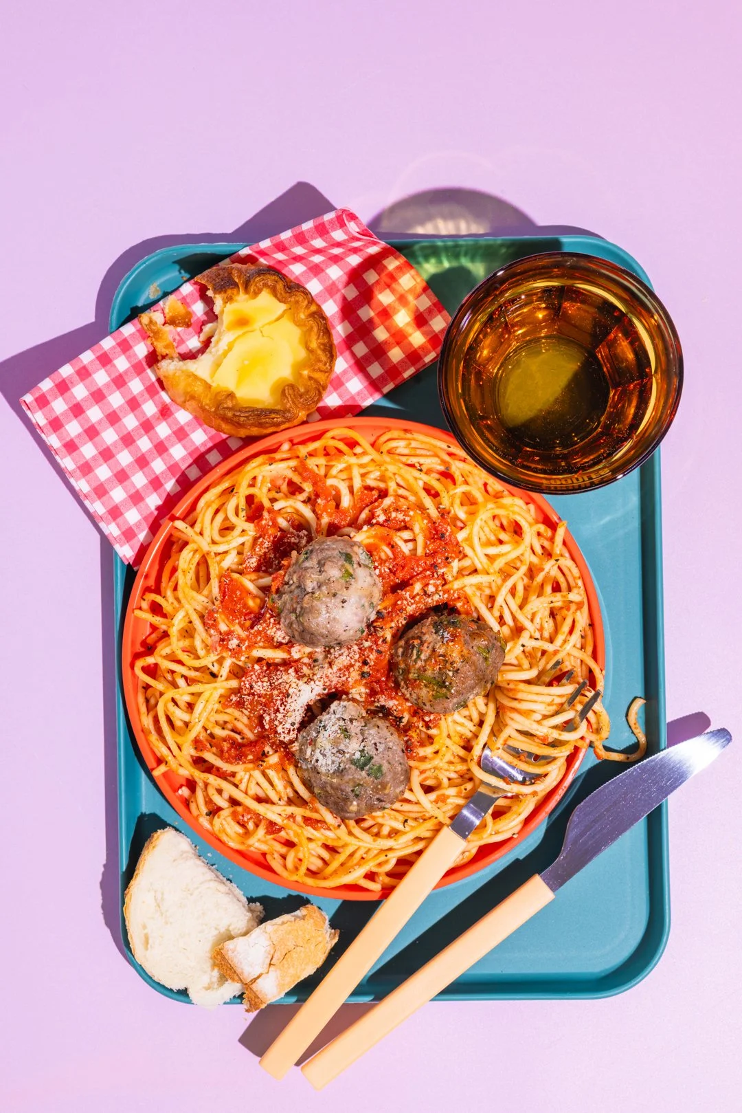 Assiette de spaghetti avec trois boulettes de viande, fromage râpé, pain, une tartelette aux œufs, une verre de boisson et des couverts sur une assiette.