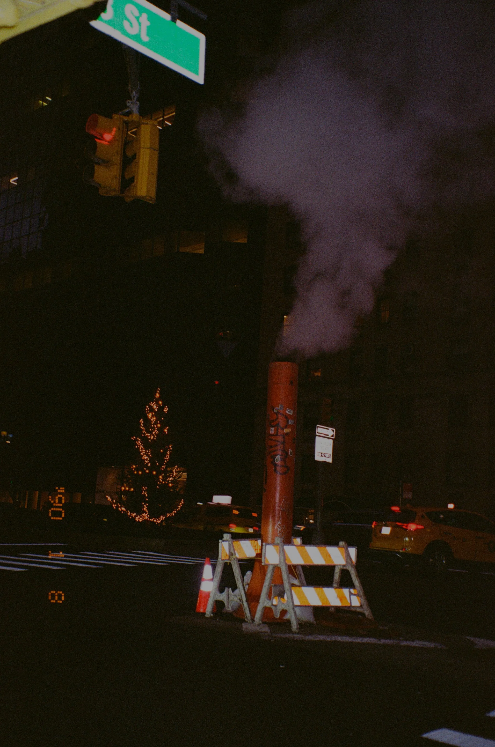 Fumée s'échappant d'une cheminée industrielle dans un environnement urbain nocturne, avec des voitures et un arbre décoré de lumières en arrière-plan.