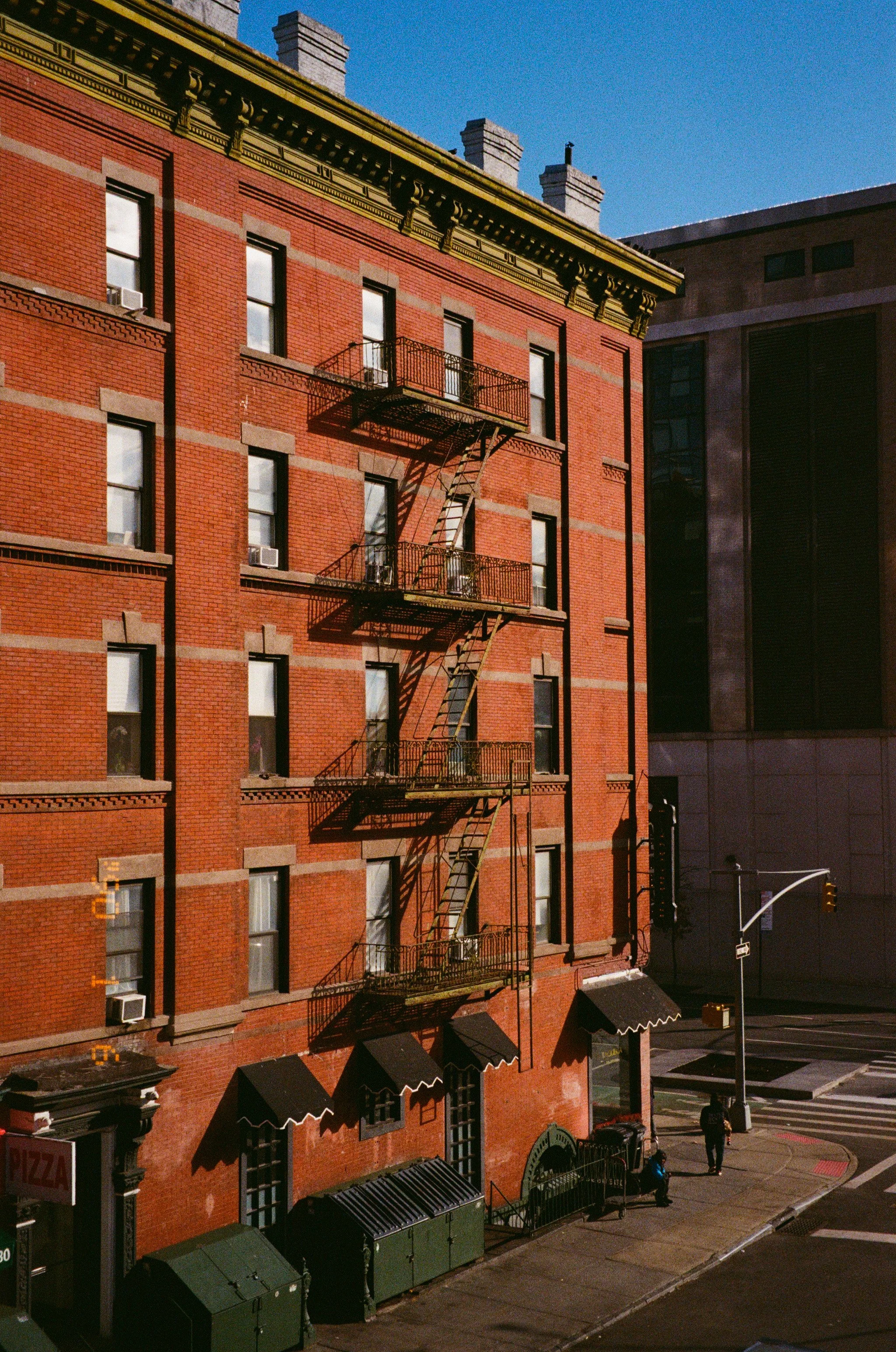 Un bâtiment en briques rouges avec des fenêtres et des balcons de feu en fer. Il y a des poubelles et deux personnes marchant sur le trottoir en face, avec un feu de signalisation à côté.