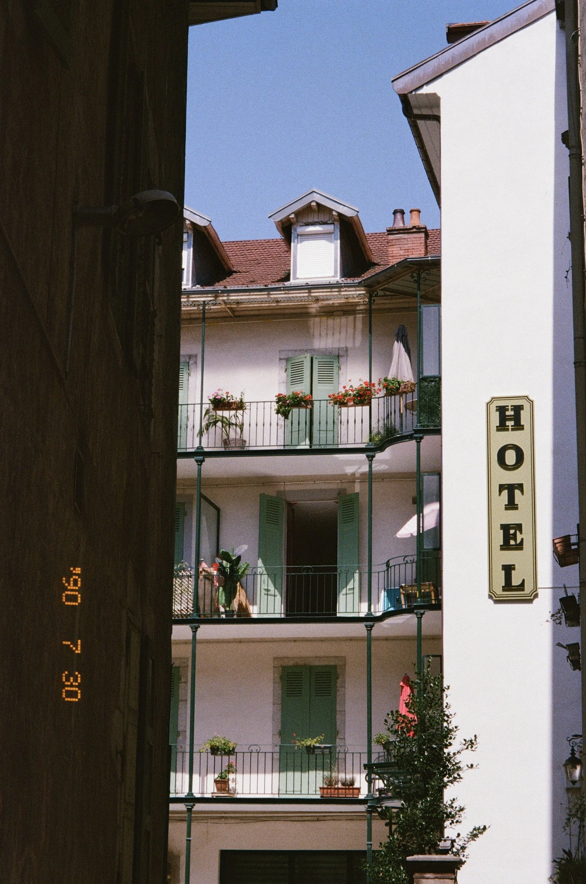 Vue d'un bâtiment d'hôtel à plusieurs étages avec des balcons décorés de fleurs, vue du côté d'une ruelle étroite sous un ciel bleu clair.