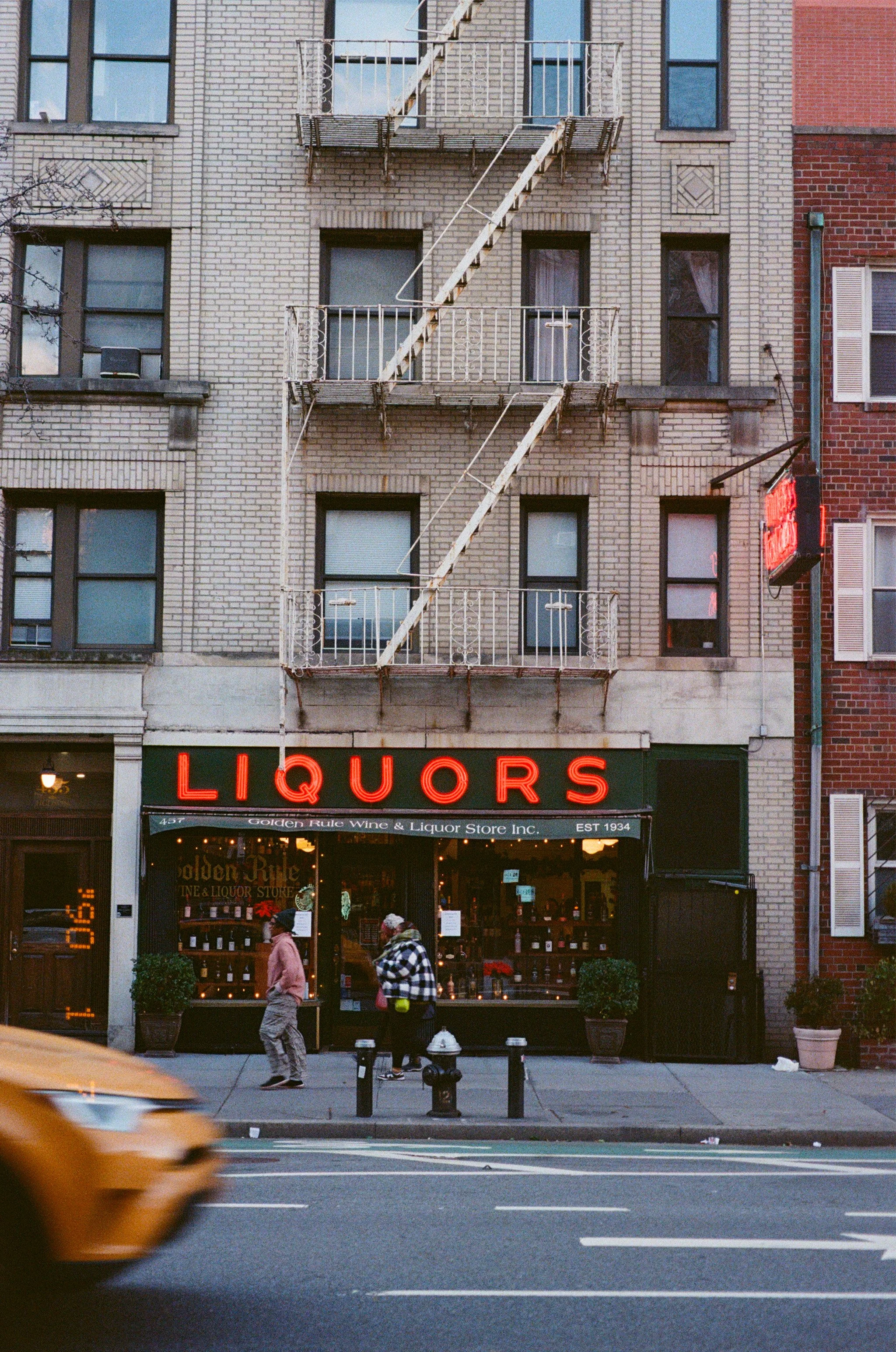 Façade d'un magasin de spiritueux avec un grand panneau néon 'LIQUORS' et deux personnes marchant sur le trottoir devant. La façade est en briques et comporte un balcon en fer forgé au premier étage, avec un bâtiment en arrière-plan.