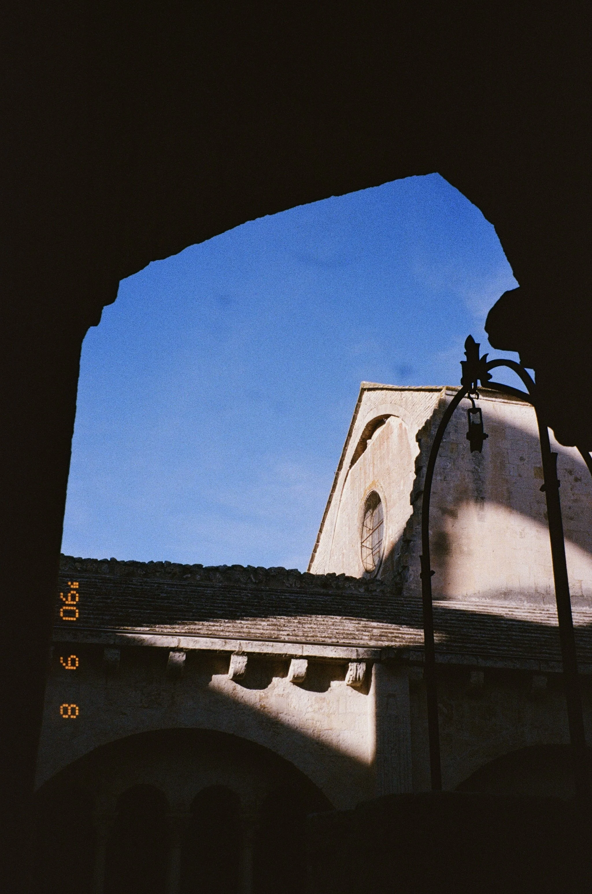 Vue from inside un cloître montrant une église en pierre sous un ciel bleu, avec une lampe en métal noire à l'avant-plan.