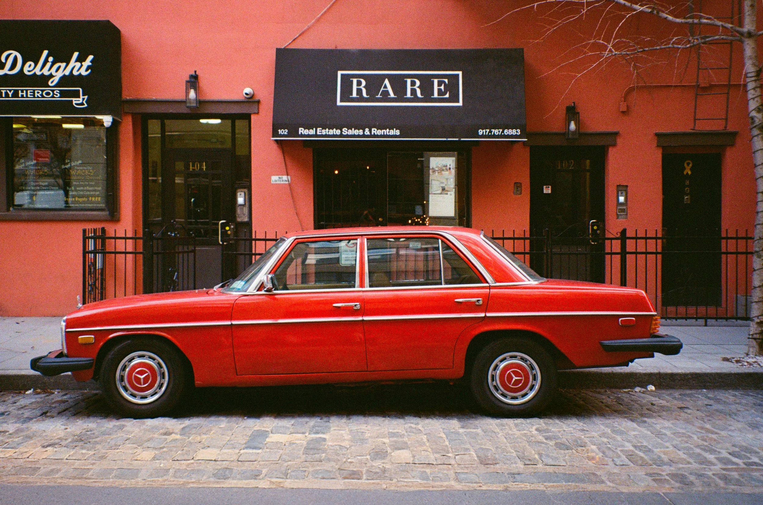 Voiture rouge classique stationnée sur un trottoir, devant un bâtiment orange avec des enseignes en anglais, y compris une qui dit RARE. L'arrière-plan montre une fenêtre et une porte, avec une branche d'arbre à droite.