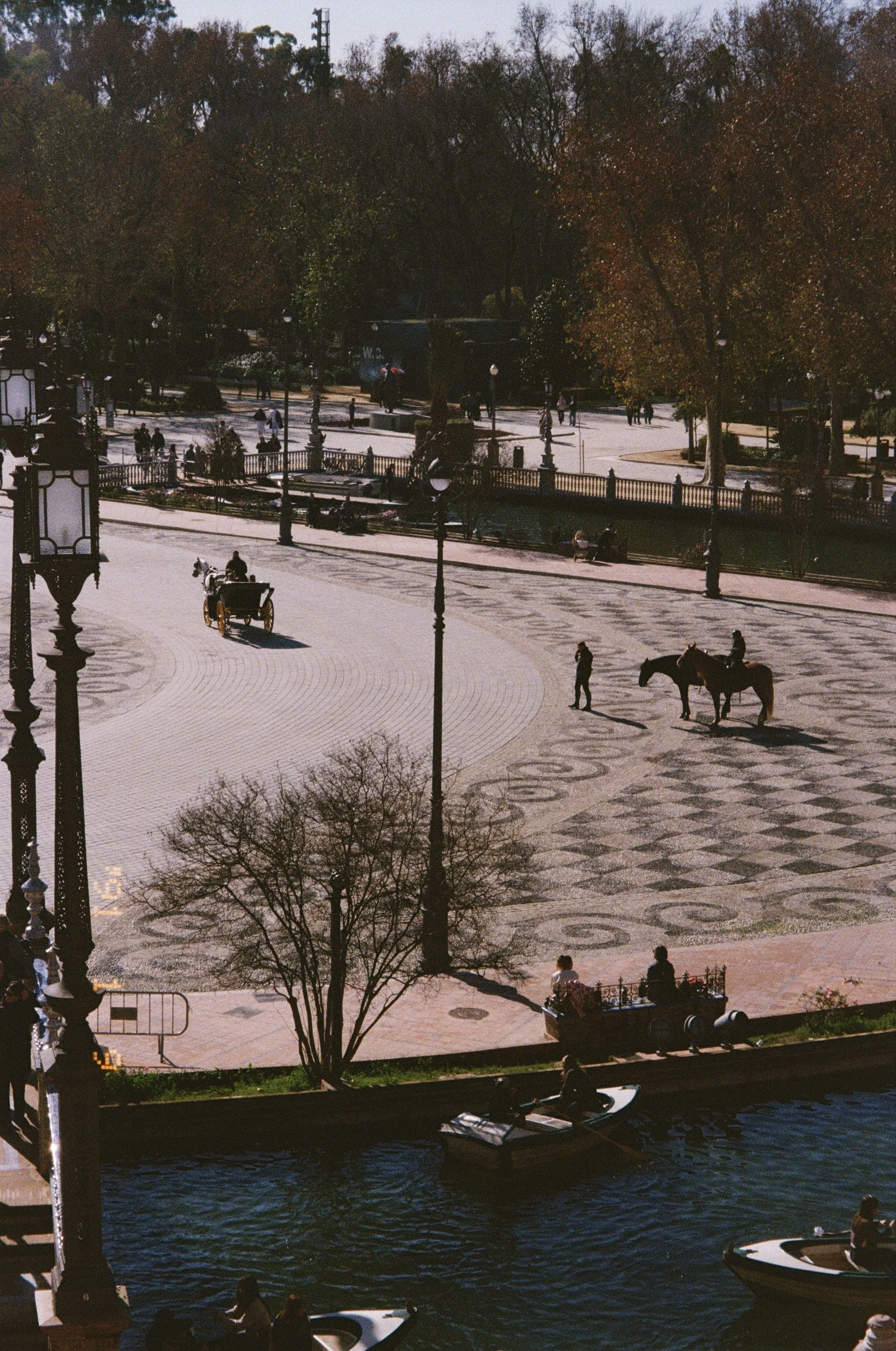 Une place publique pavée, avec des lampadaires, des arbres, des personnes assises, un cheval avec un cavalière, une calèche, et un canal avec des petits bateaux, en plein jour.