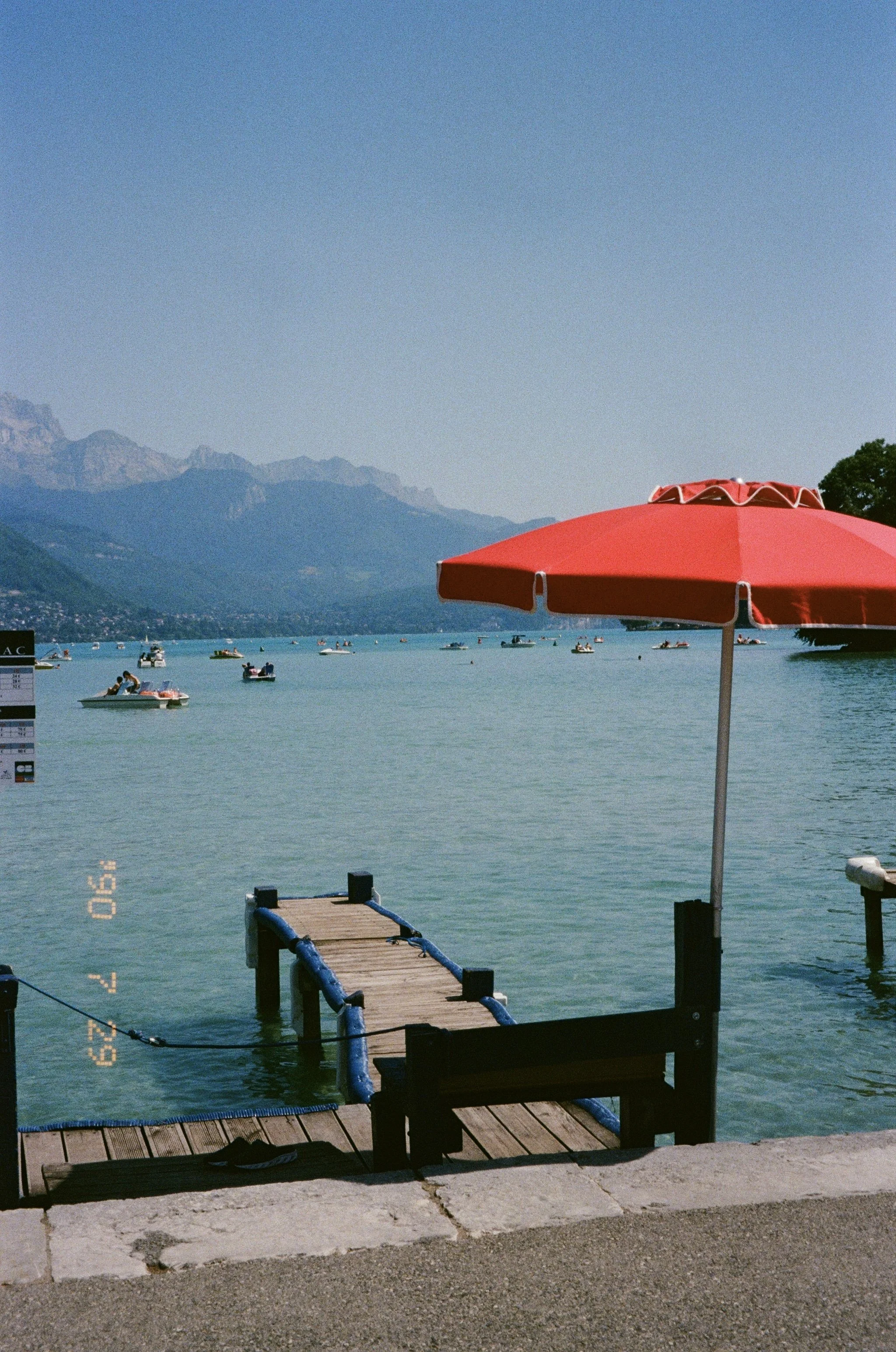 Un quai en bois avec un parasol rouge sur le bord d'un lac ou mer, avec des montagnes en arrière-plan et plusieurs bateaux flottant à la surface de l'eau.