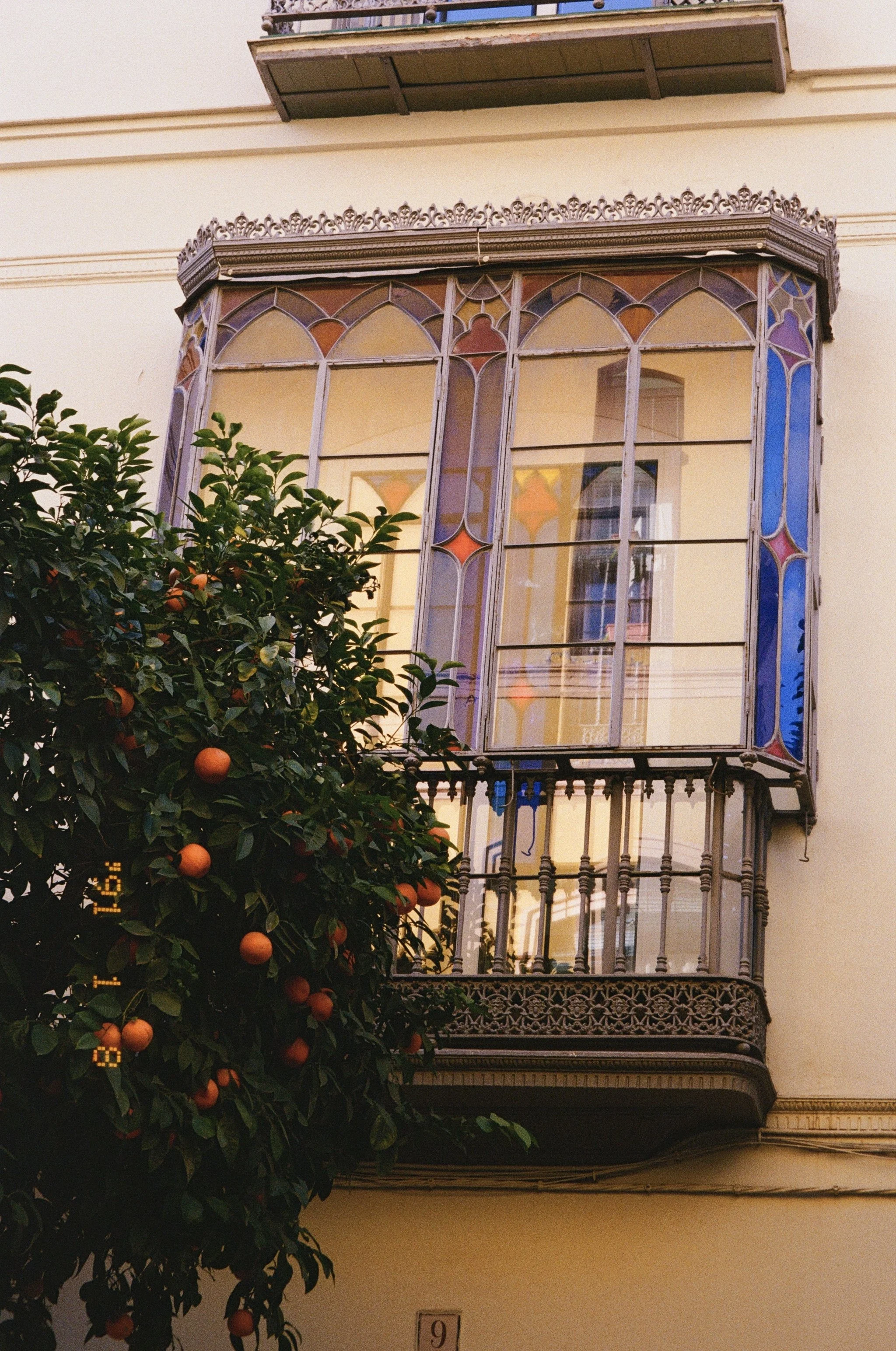 Fenêtre en vitrail colorée avec balcon en fer forgé sur un bâtiment de style classique, avec un arbre fruitier dans le premier plan.