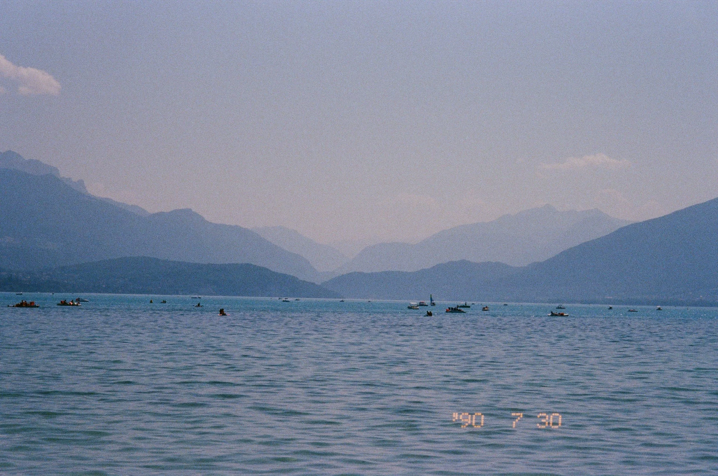 Une vue d'un lac avec plusieurs bateaux à voile et en moteur, entouré de montagnes là-bas, sous un ciel clair et ensoleillé.