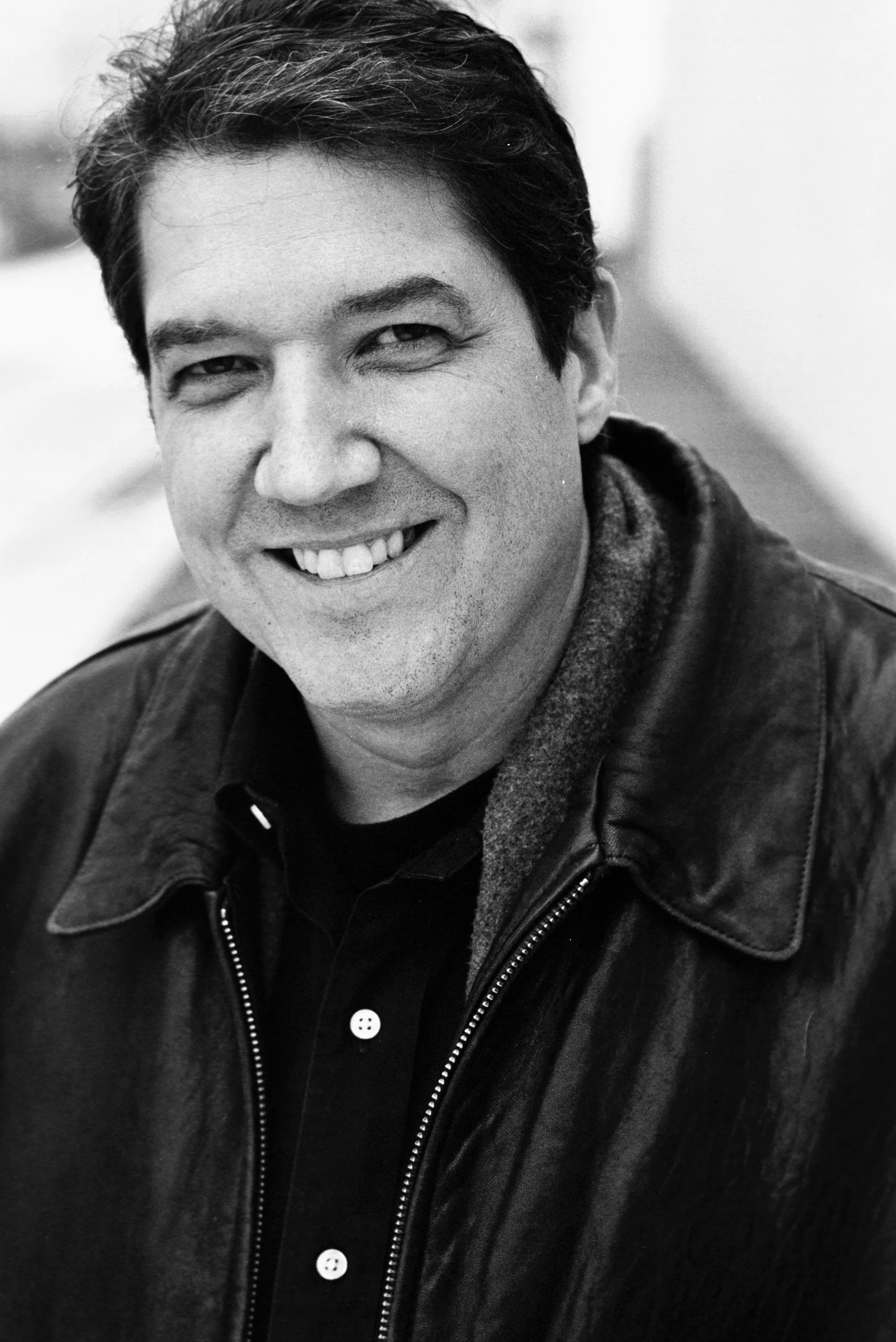 Author Michael Dimercurio smiling with dark, slightly tousled hair, wearing a dark leather jacket and a dark shirt with buttons.