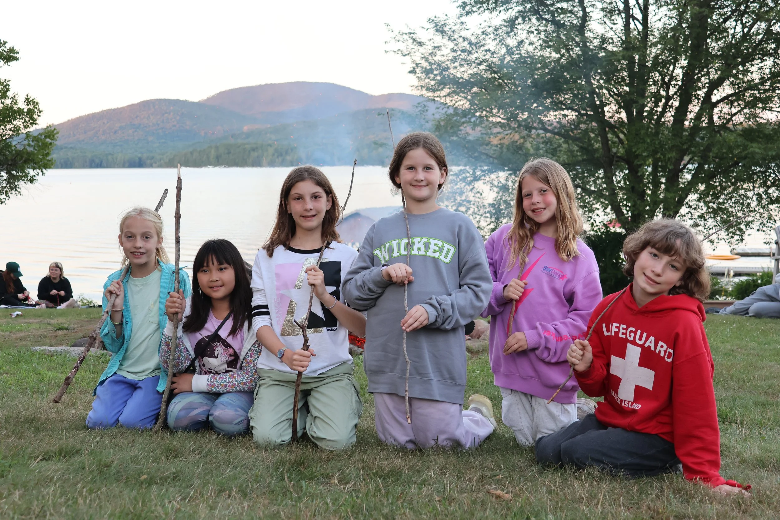 Group of children outdoors near lake, holding sticks, with mountains, trees, and other people in the background during daytime.