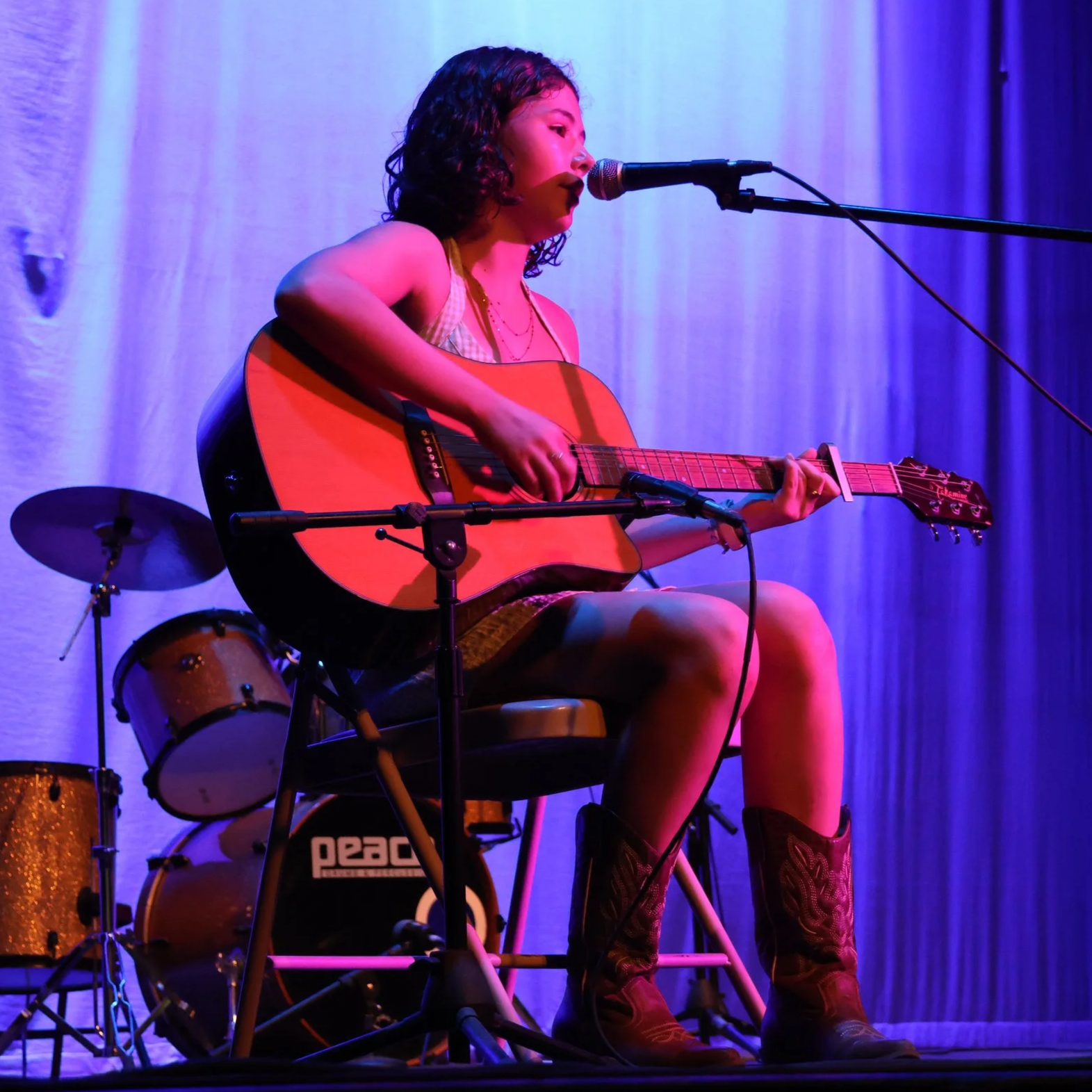 Young girl with curly hair singing into a microphone and playing an acoustic guitar on stage with blue and purple lighting, and a drum set in the background.