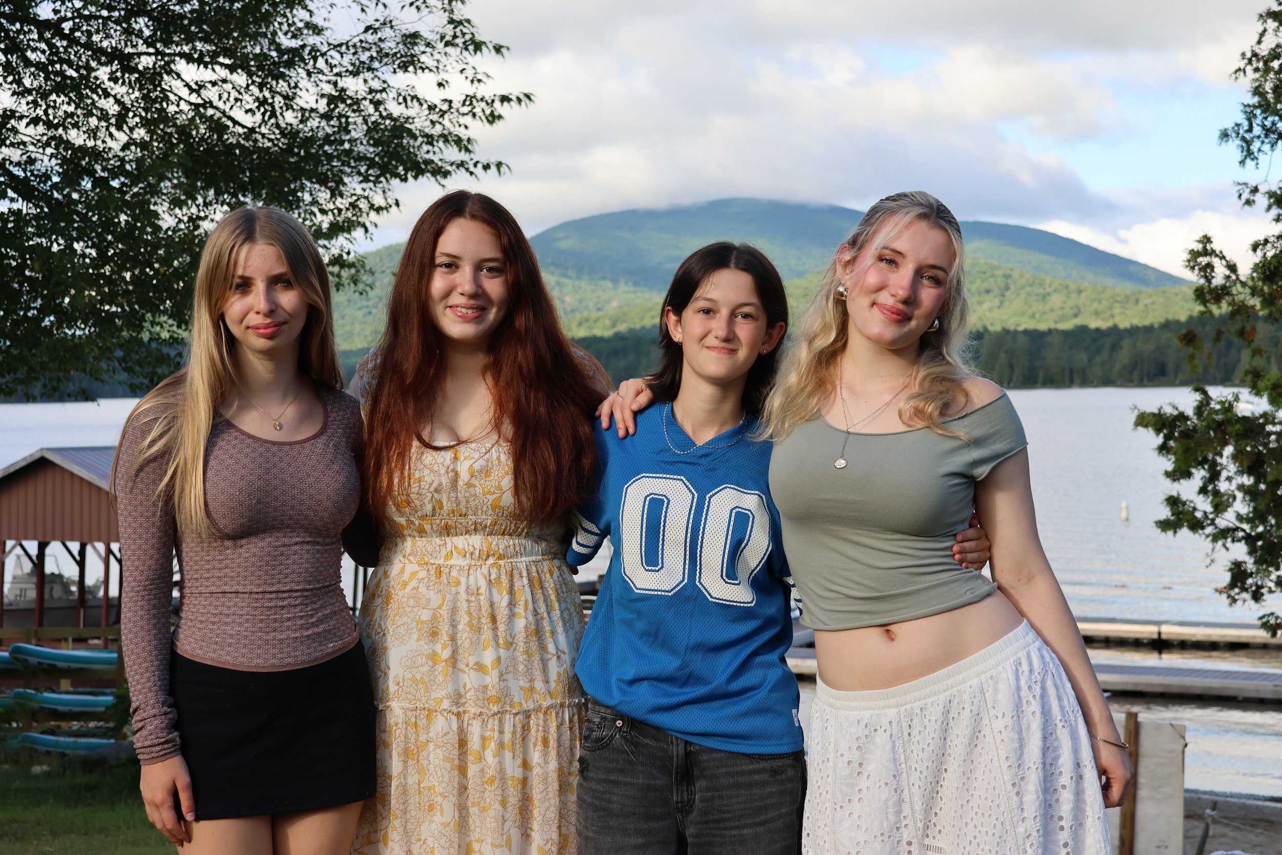 Four young women standing together outdoors near a lake with mountains in the background. They are smiling and have their arms around each other, dressed casually.